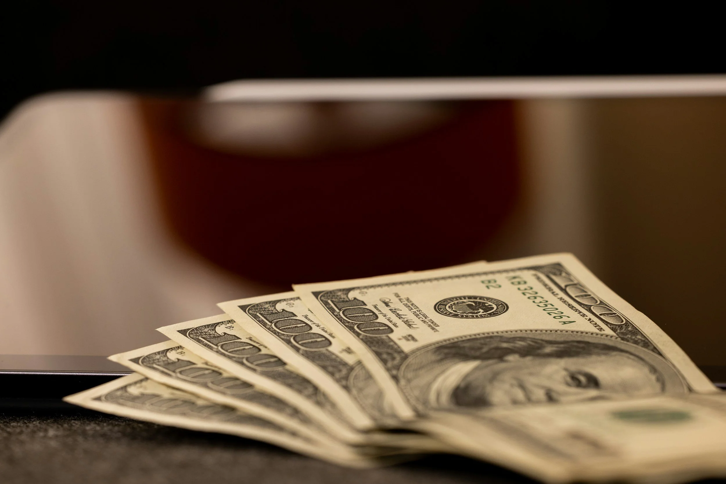 Four 100-dollar bills partially fanned out on a dark surface with a reflective, metallic object behind them.