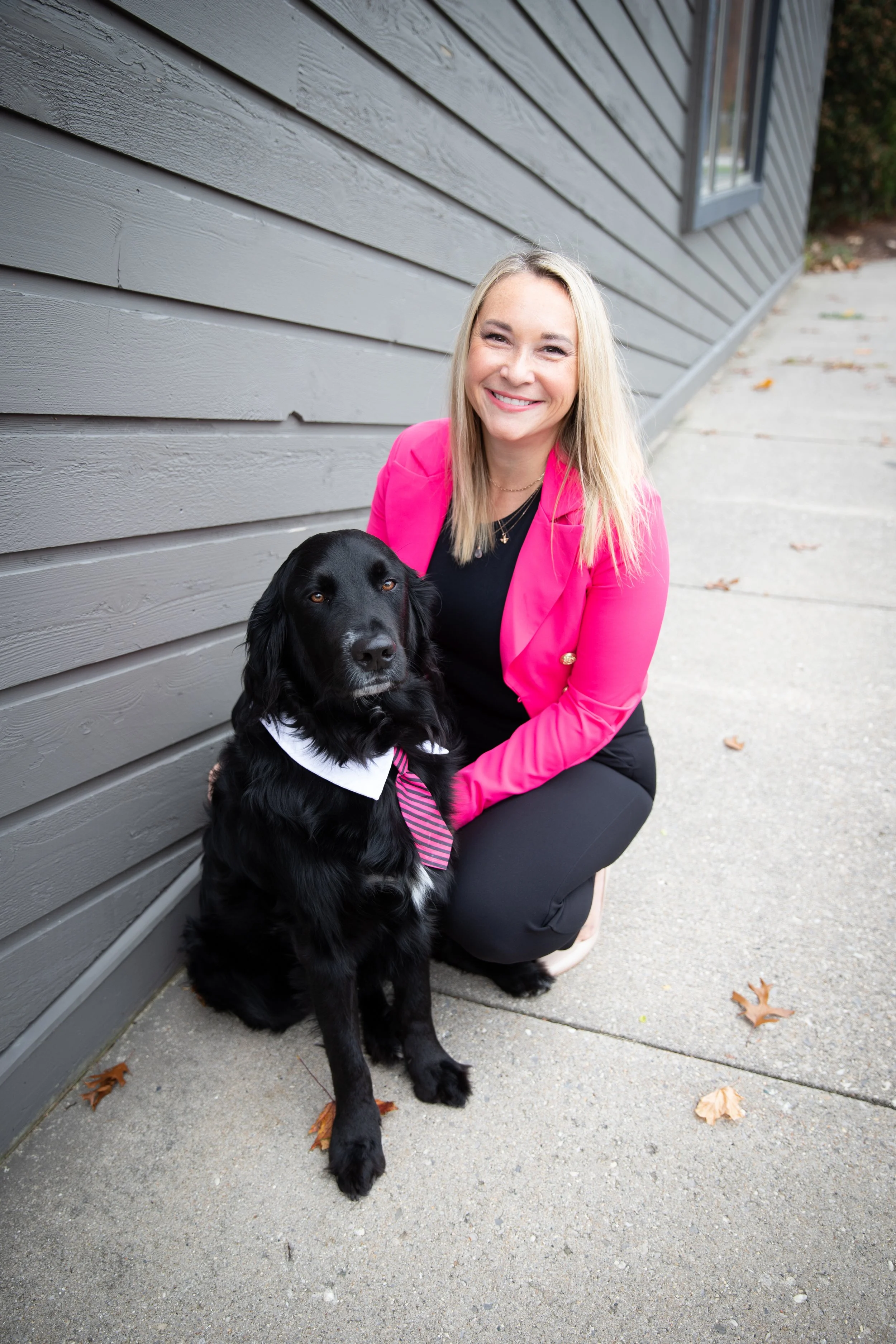 A woman in a pink blazer squatting next to a black dog wearing a white collar and a pink and gray striped tie, outdoors near a gray wooden wall.