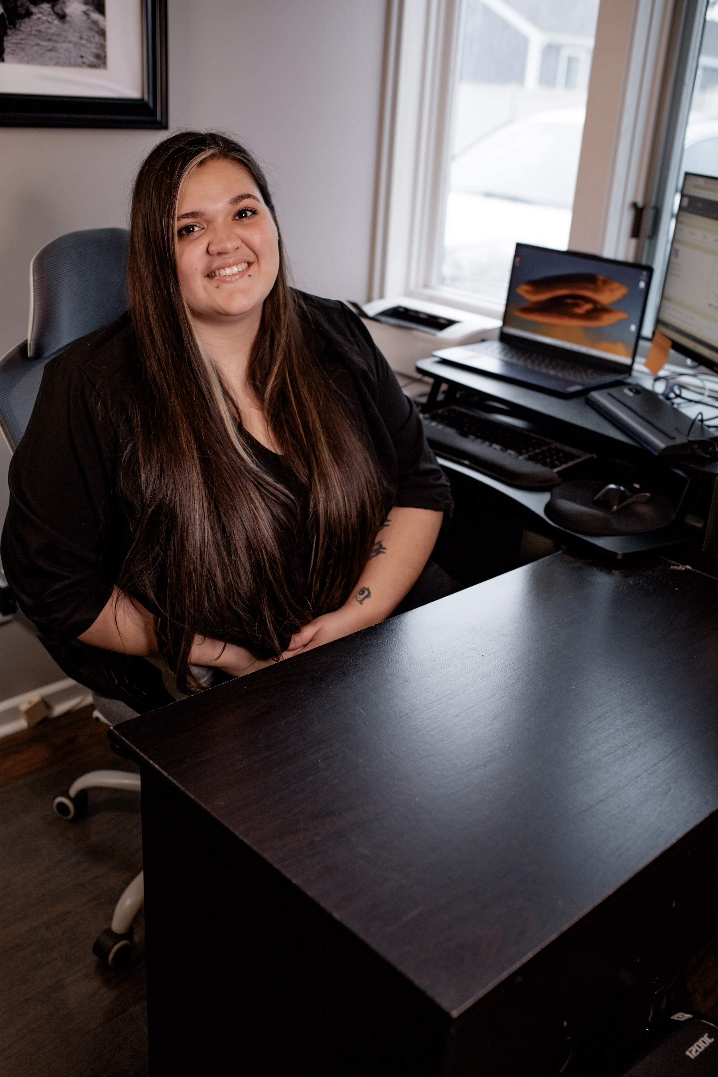 A woman with long brown hair and tattoos on her arm sitting at a black desk in an office, smiling at the camera. There are two computer monitors, one showing a desktop background and the other displaying an email or document, and a laptop on the desk near a window.