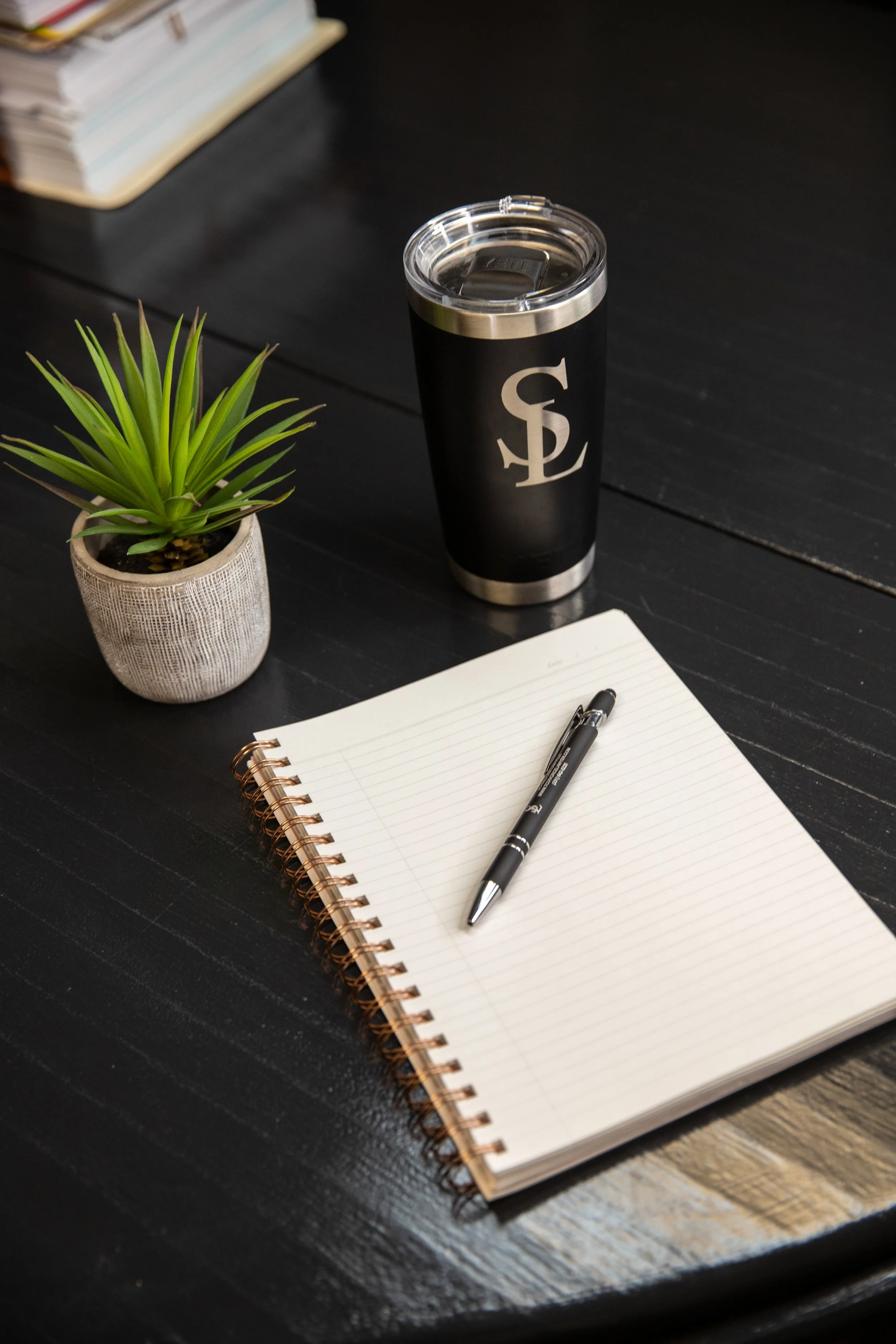A black table with a spiral notebook, a black pen, a potted green succulent, a stainless steel travel mug with a black sleeve and logo, and a stack of books in the background.
