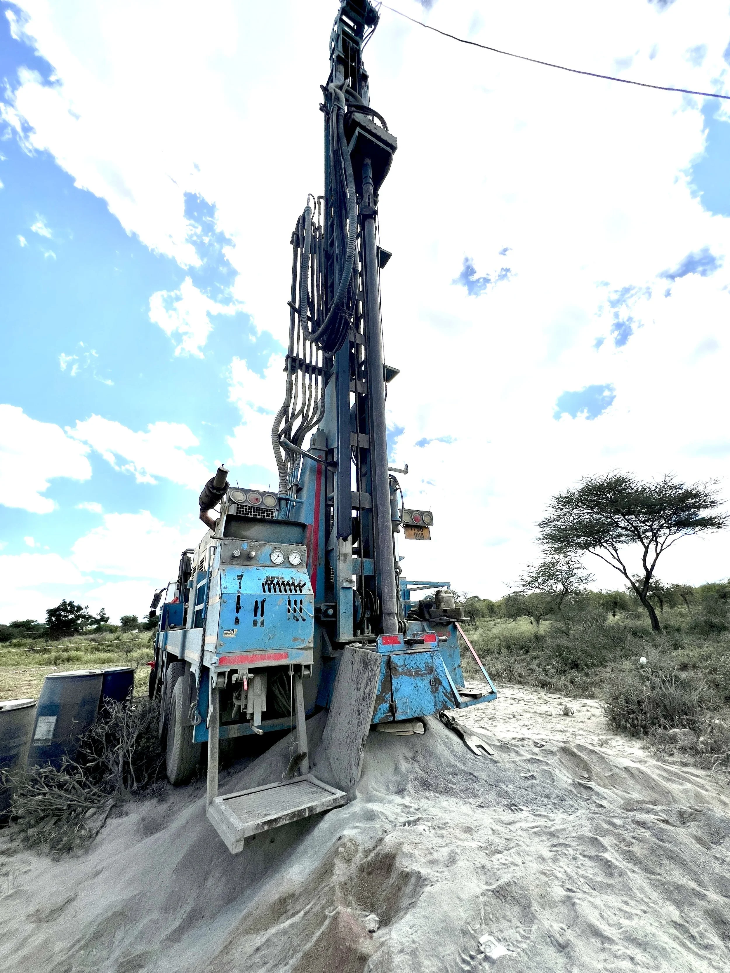 A tall drilling rig machine in a dry, semi-arid landscape with sparse vegetation and a few trees, under a partly cloudy sky.