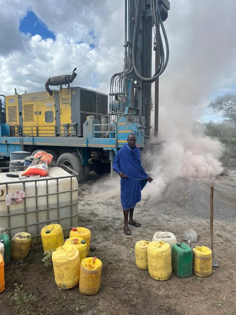 A man in blue traditional Maasai clothing standing next to a drilling rig on a dry, dusty land. There are yellow and green water containers nearby, with some on the ground and others on a cart. Dust and steam are visible around the drilling equipment, and a cloudy sky is overhead.