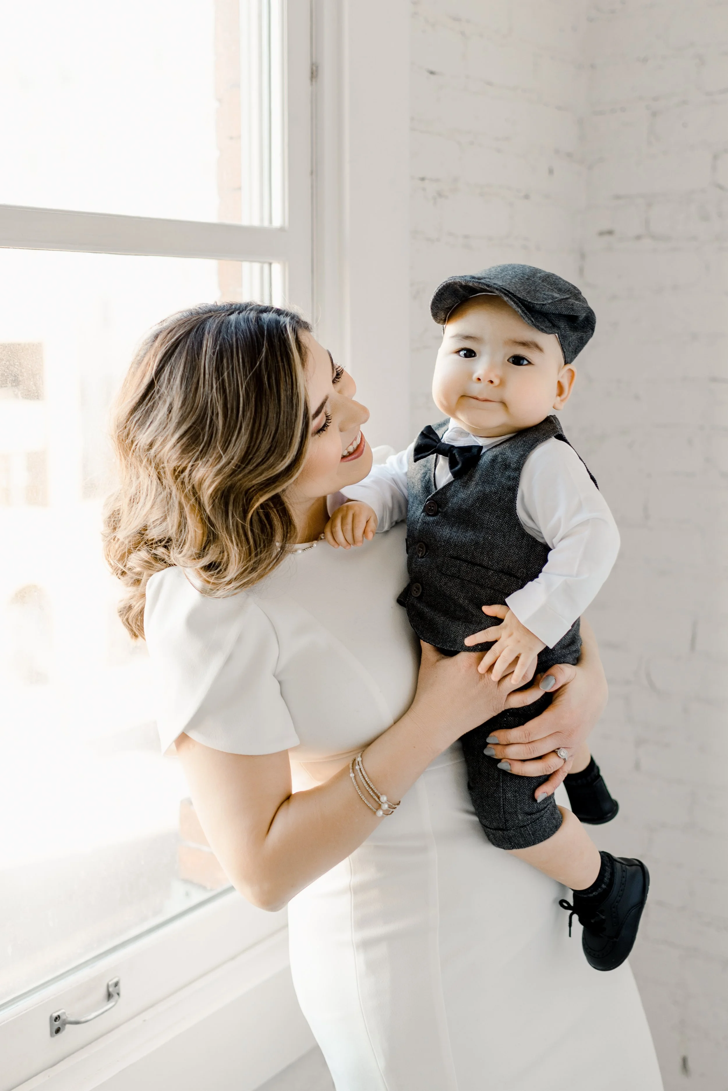 A woman smiling and holding a young boy dressed in vintage attire near a window.