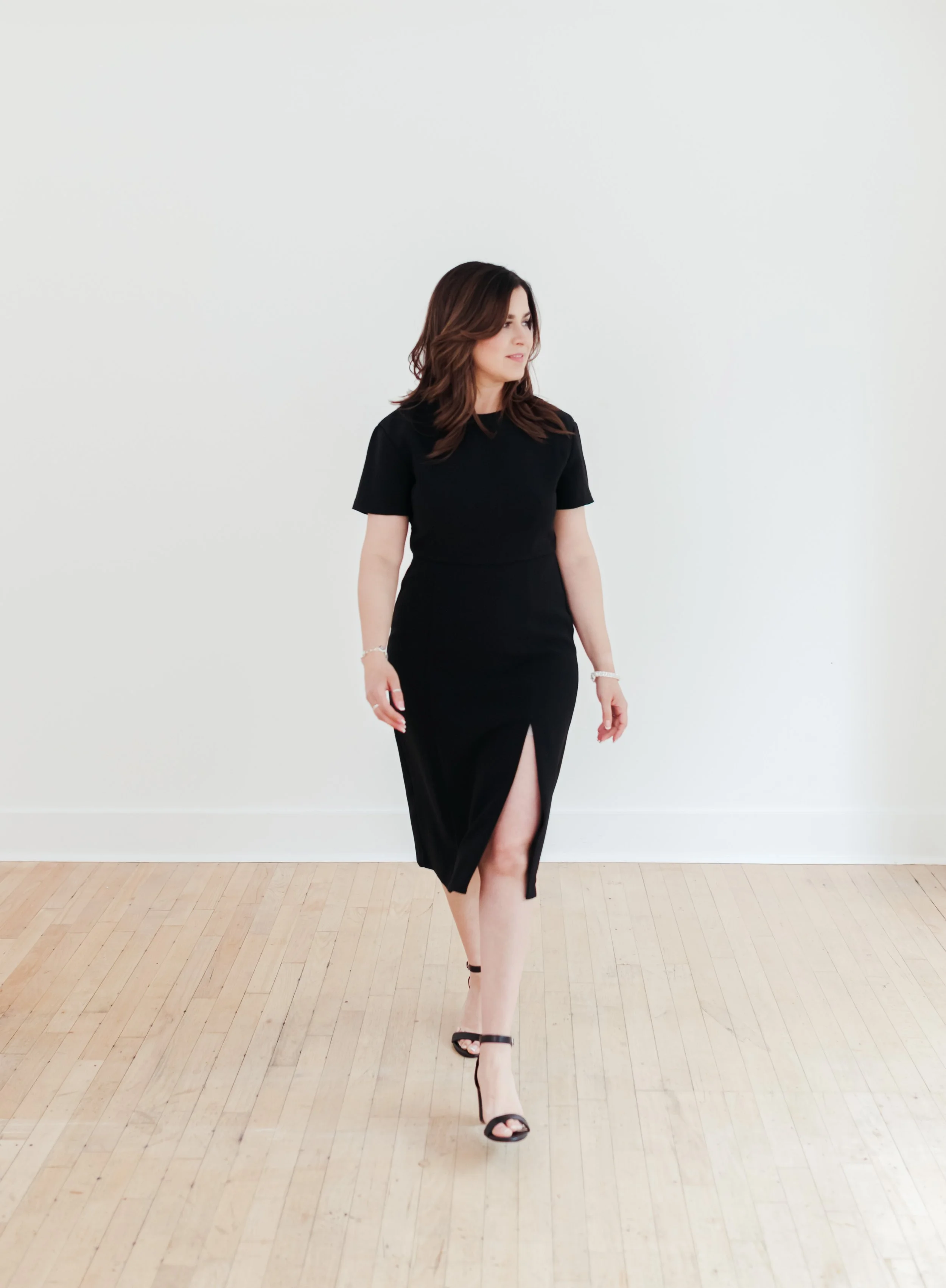 A woman in a black dress walking on a wooden floor against a plain white wall.