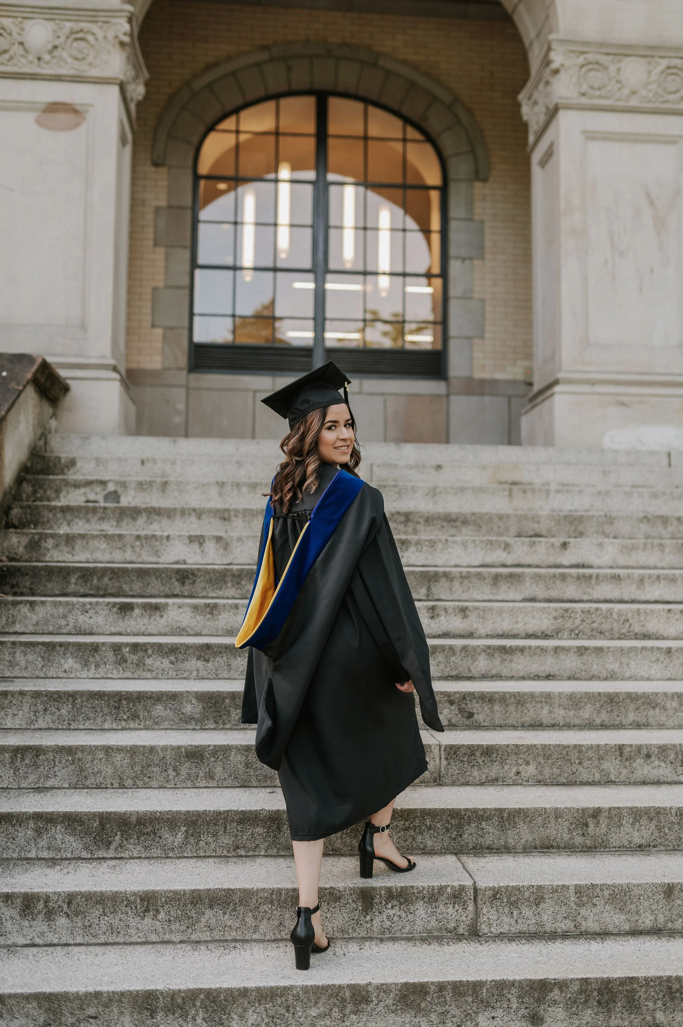 A woman in a graduation cap and gown standing on the stairs outside a building, smiling at the camera.