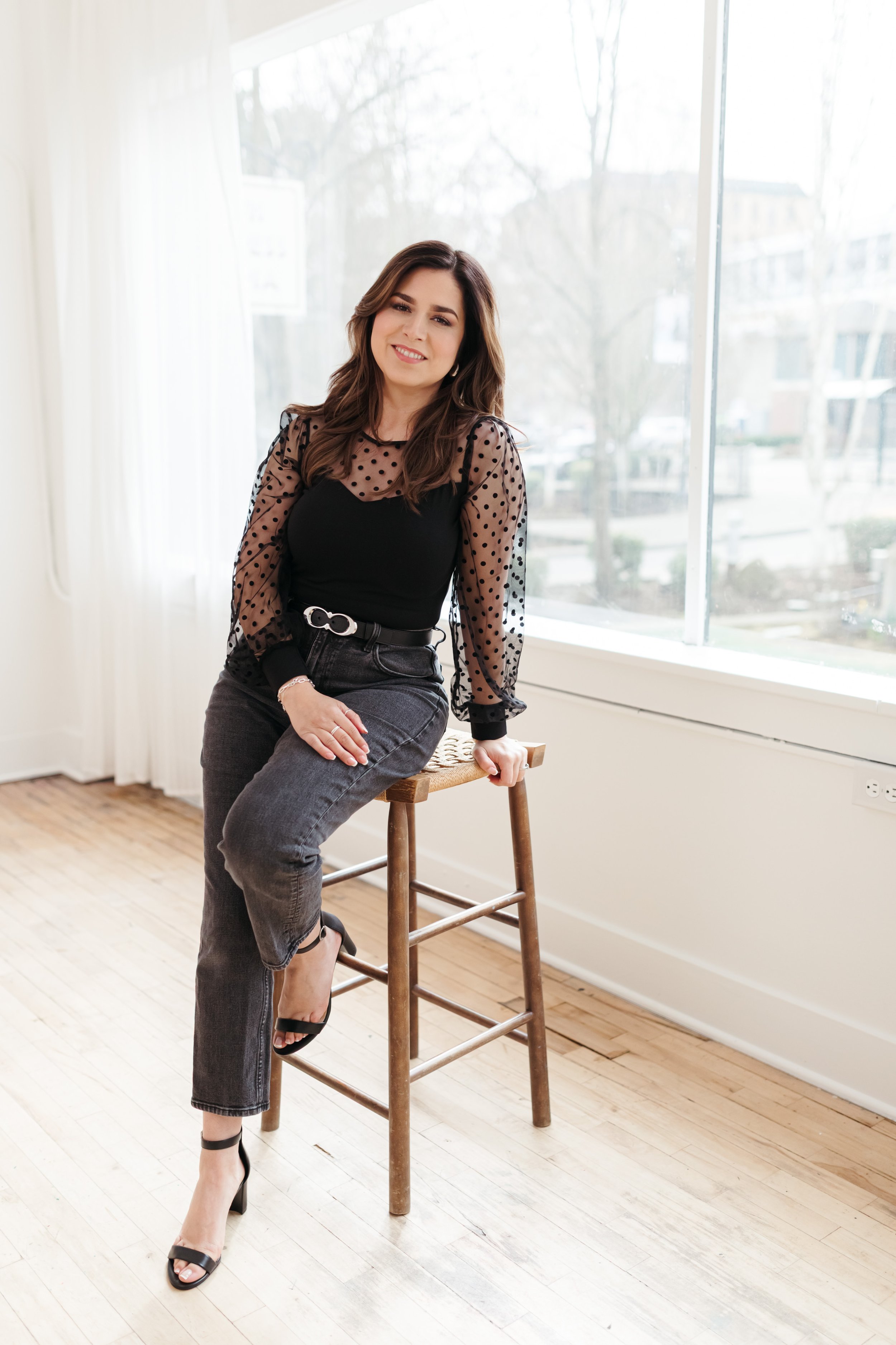 A woman sitting on a high wooden stool near a large window, smiling, wearing a black top with sheer polka dot sleeves, gray jeans, and black high heels.