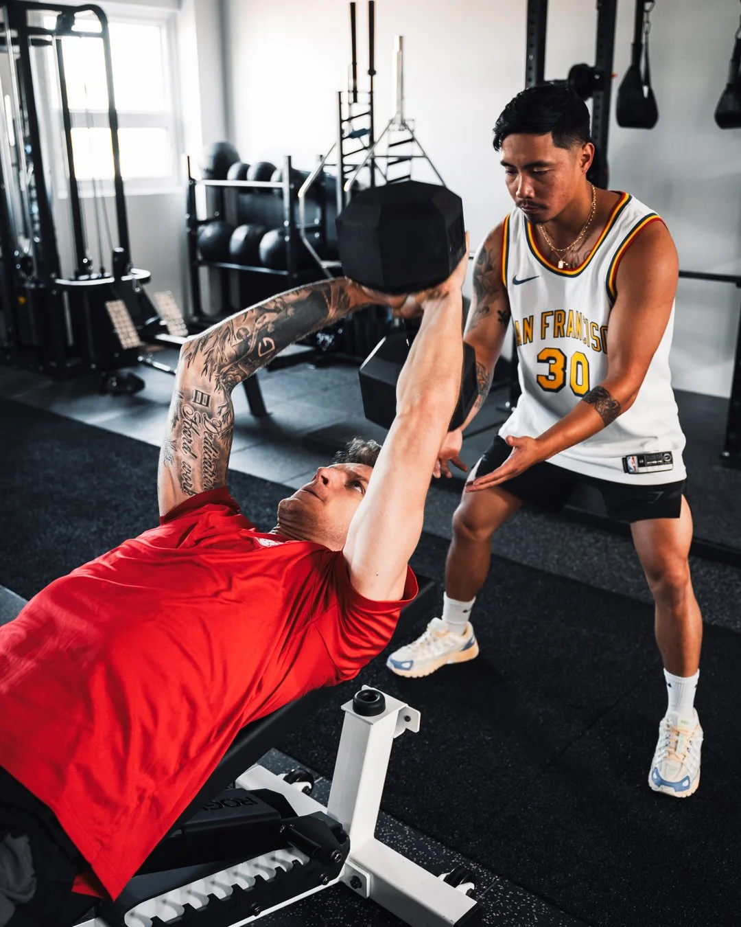 A man in a red shirt lying on a bench performing a dumbbell chest press exercise with a trainer spotting him in a gym.