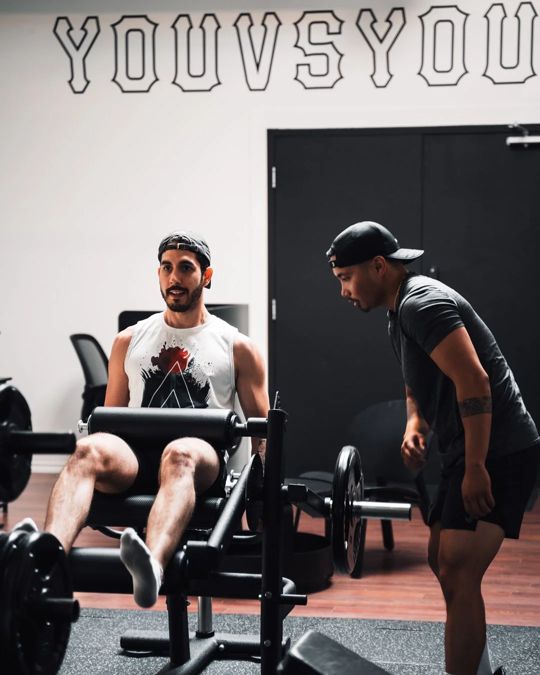 Two men at the gym, one sitting on a leg curl machine and the other standing next to him, talking. The man seated wears a sleeveless shirt, black shorts, and a cap backward. The man standing wears a black cap, dark shirt, and shorts. The gym background has a motivational quote on the wall, partly visible.