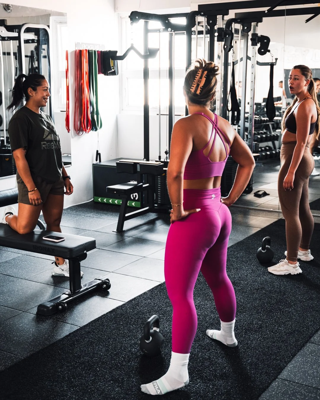 Three women in workout attire talking in a gym with fitness equipment. One woman in a black T-shirt and shorts is kneeling beside a bench, another in pink leggings and sports bra is standing with her hands on her hips, and the third in beige leggings and a black sports bra is standing nearby.