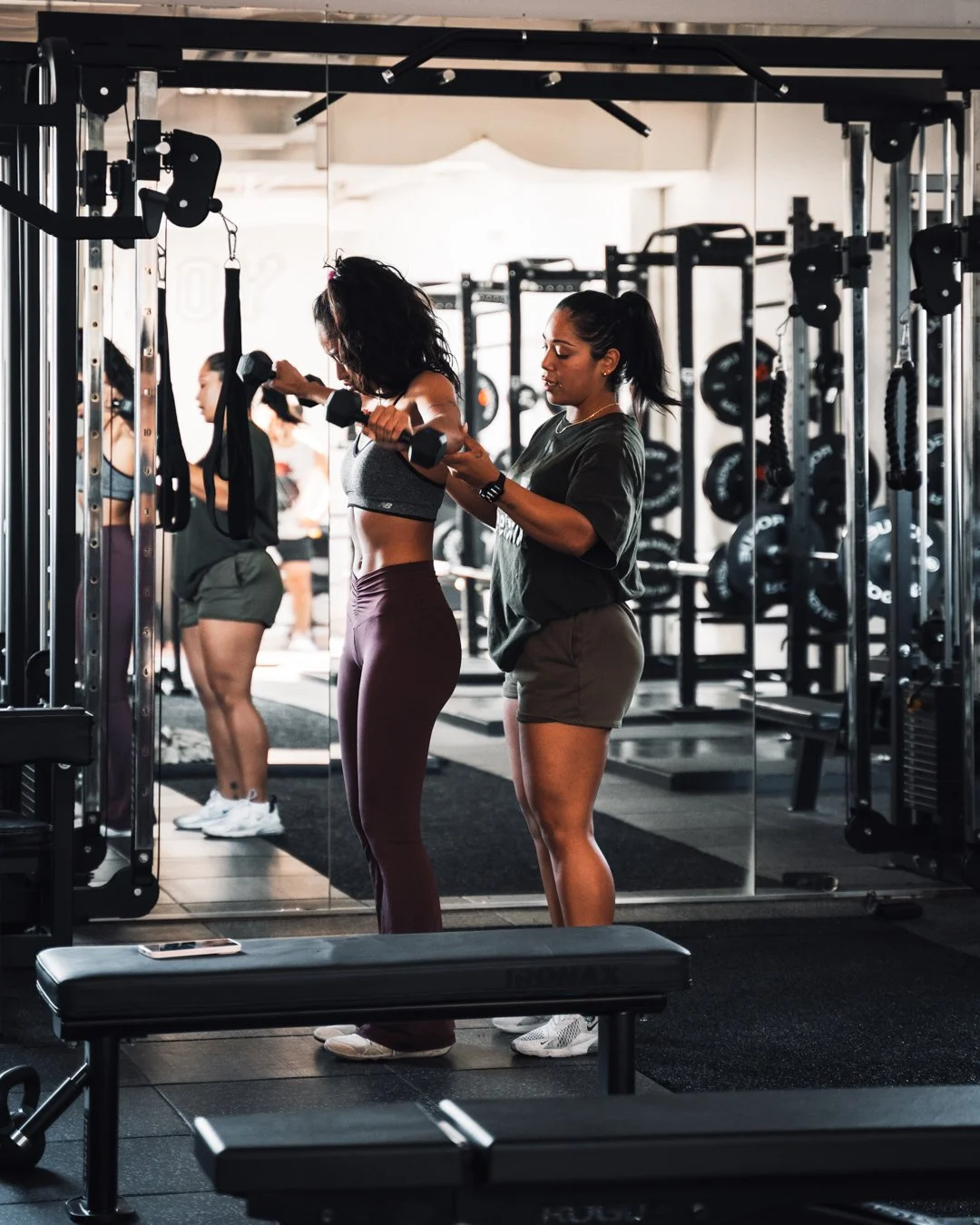 A woman receiving assistance from a trainer while lifting dumbbells in a gym, with other people working out in the background.