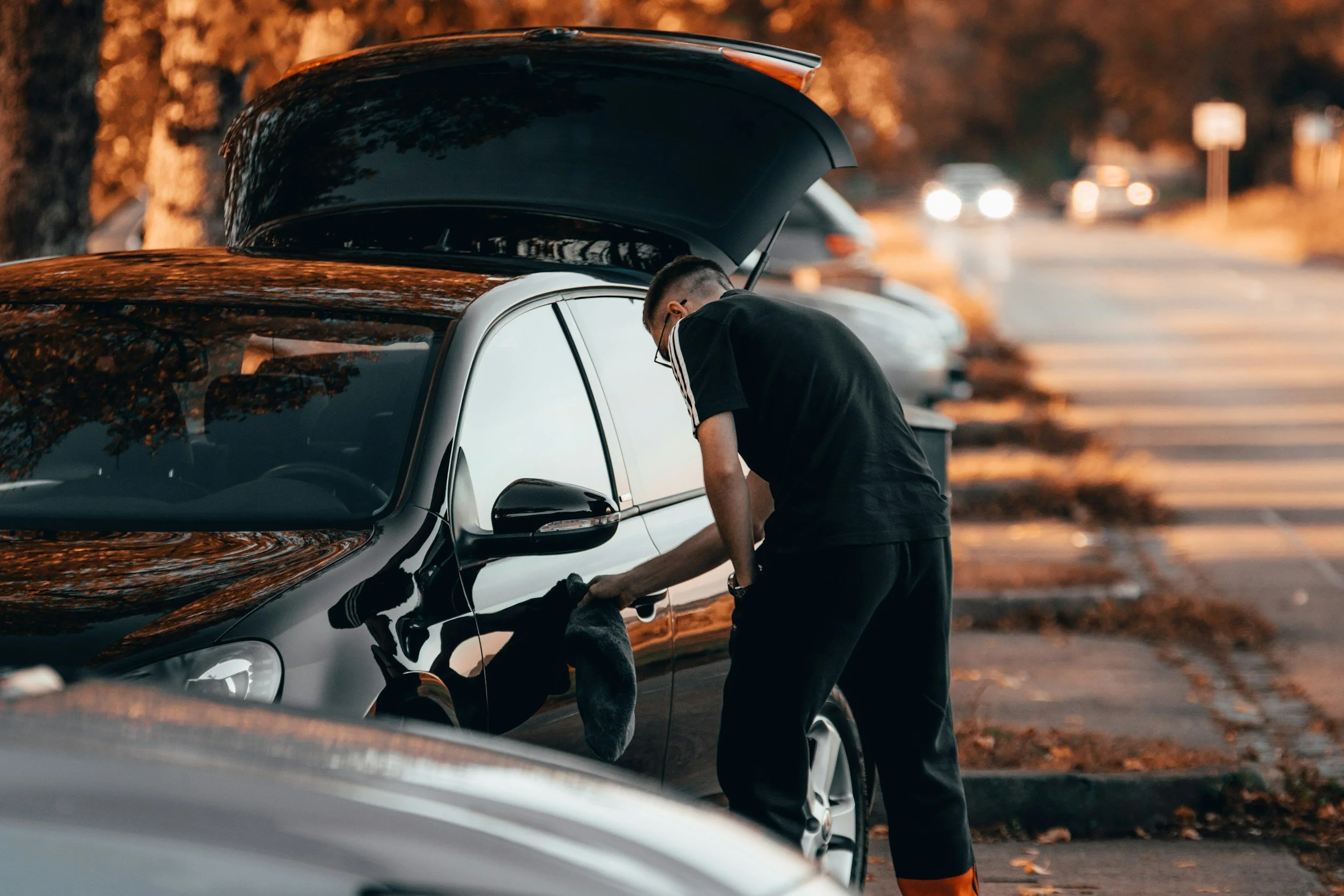 Un jeune homme charge un chien dans une voiture noire stationnée dans un parking en soirée, avec d'autres voitures en arrière-plan.