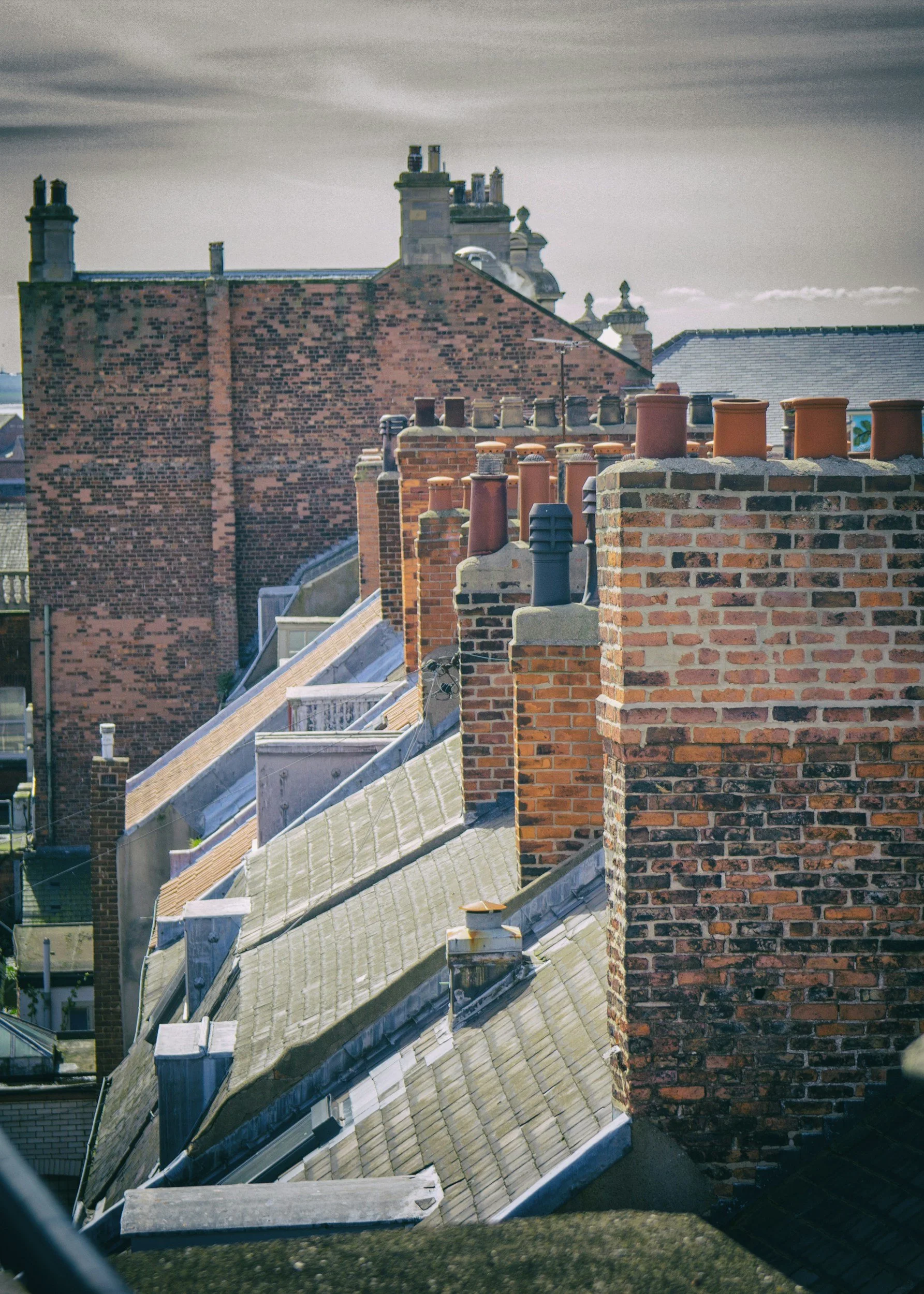 Close-up of brick chimneys and rooftops on terraced houses, with a cloudy sky in the background.