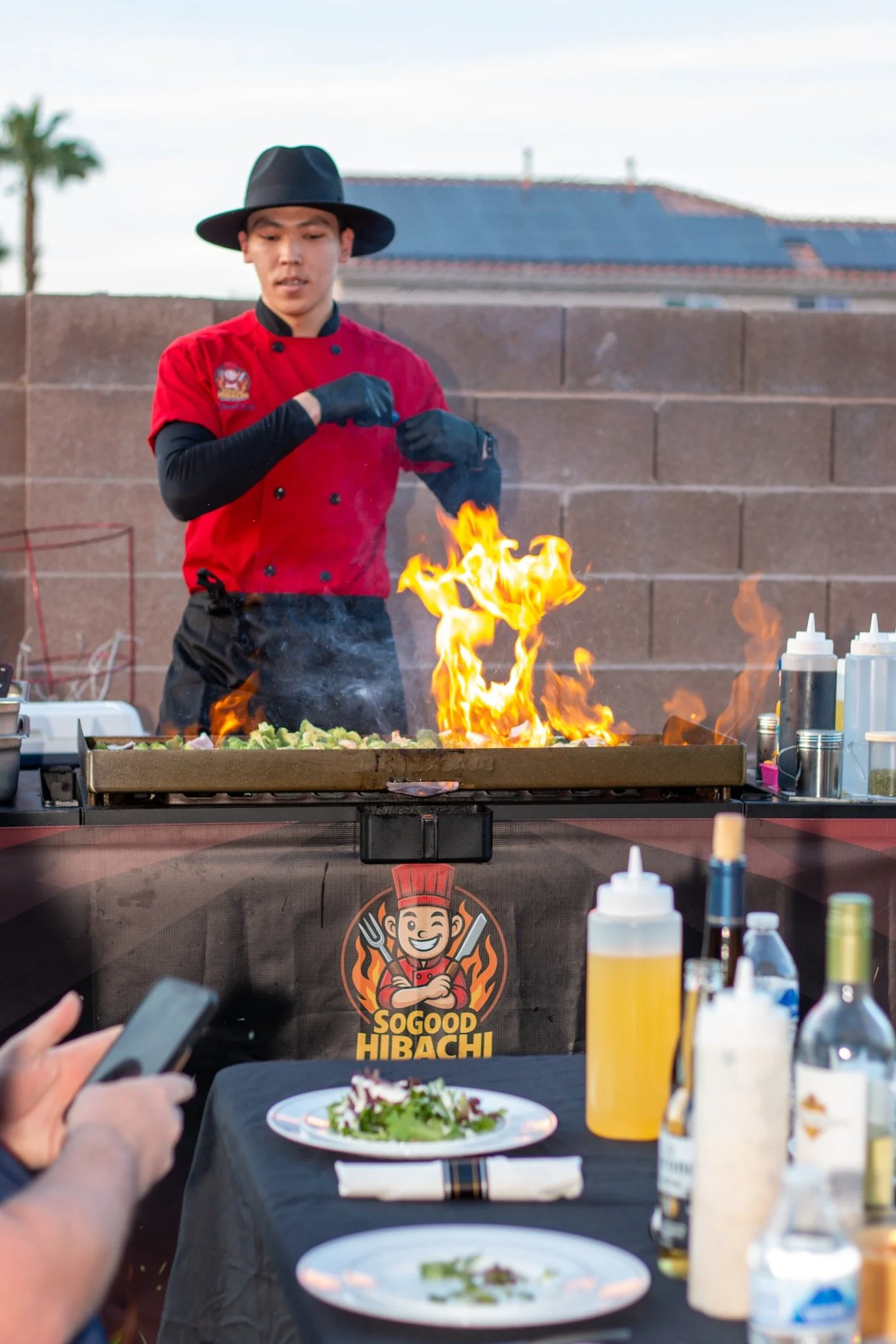 A chef wearing a red uniform and black hat stands behind a griddle with flames rising from cooking food. Outside in what appears to be a backyard, with a brick wall and house in the background, the chef is preparing food at a barbecue or street food 