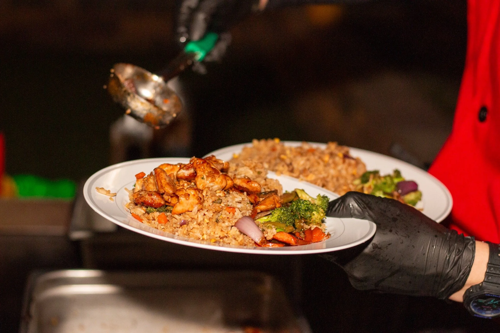 Person in black gloves serving plates of fried rice with chicken and vegetables at a buffet or catered event.
