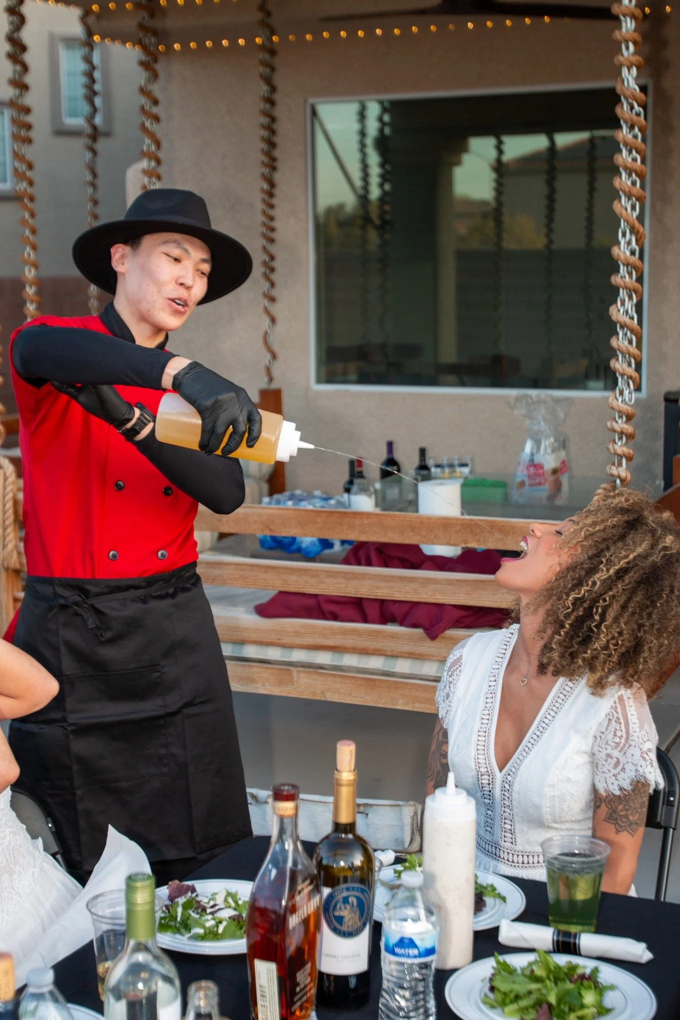 A woman with curly hair and tattoos on her arm laughing while sitting at a table, as a chef squeezes mustard on her mouth during a playful outdoor dining event.