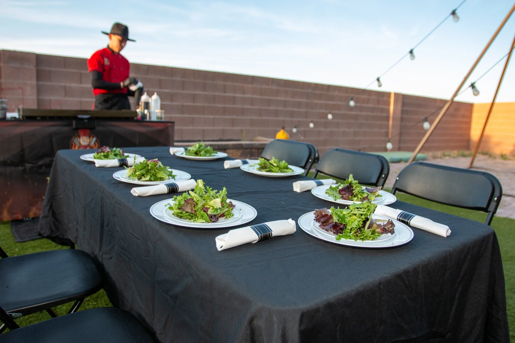 Set outdoor dining table with six plates of salad, napkins, and utensils, with a person preparing food in the background on a grill, and string lights overhead, against a brick wall.
