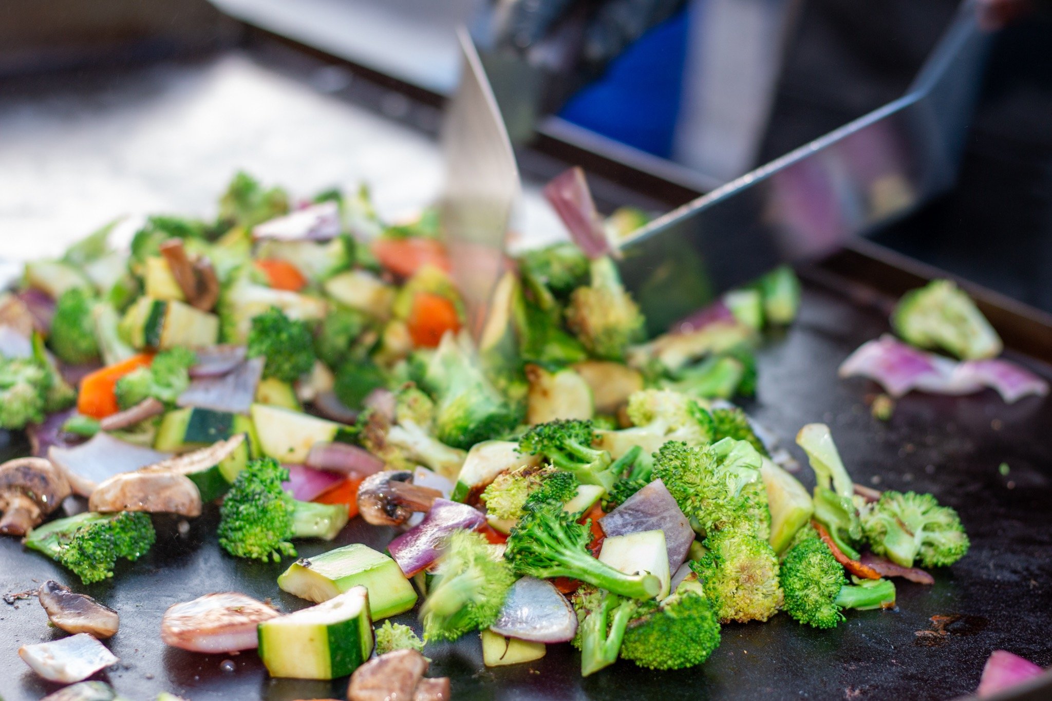 Cooked mixed vegetables, including broccoli, zucchini, red onion, and mushrooms, on a flat surface with a spatula.