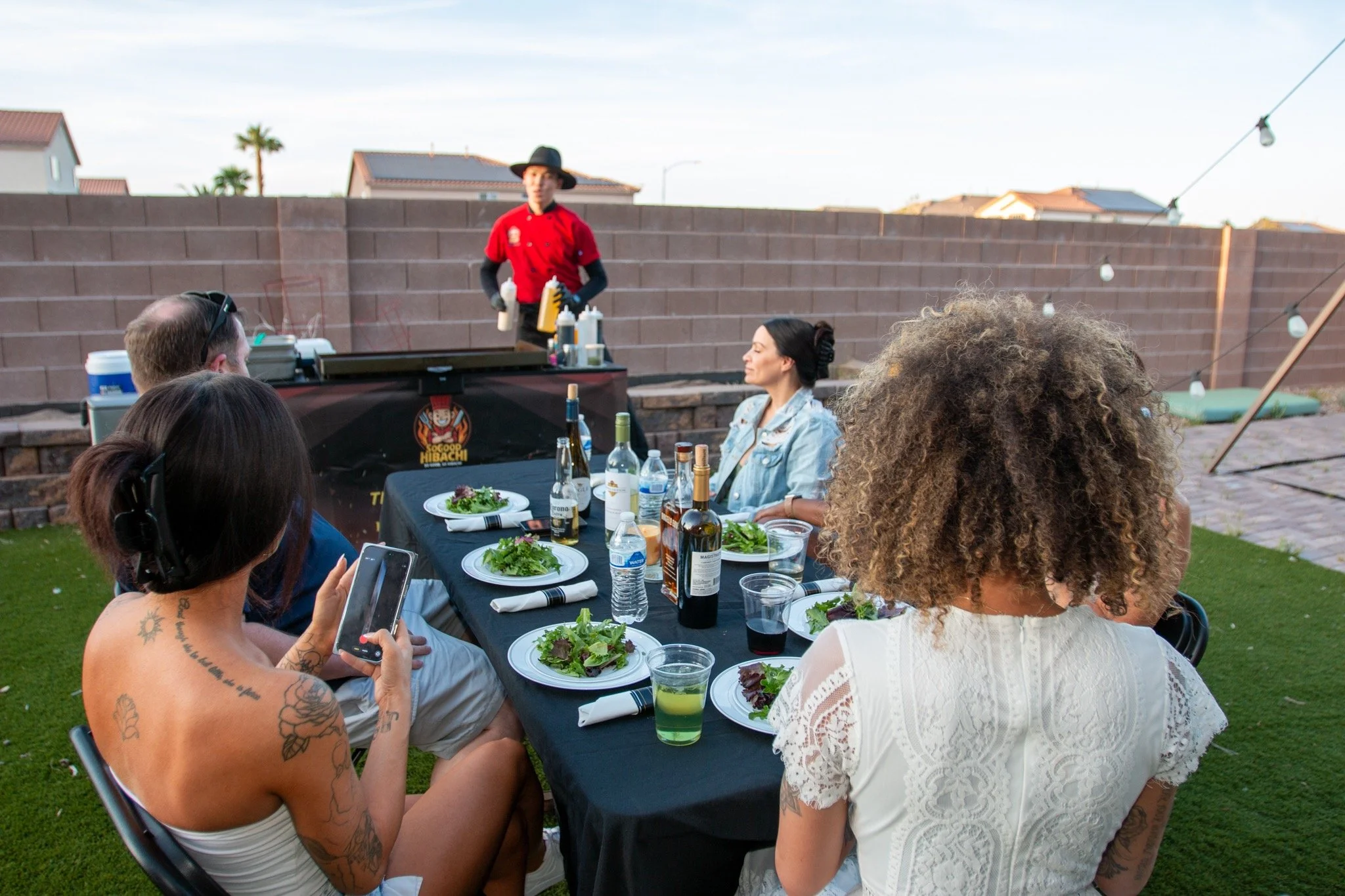 People sitting at an outdoor dinner table with food and drinks, watching a chef cooking on a grill at a backyard gathering.