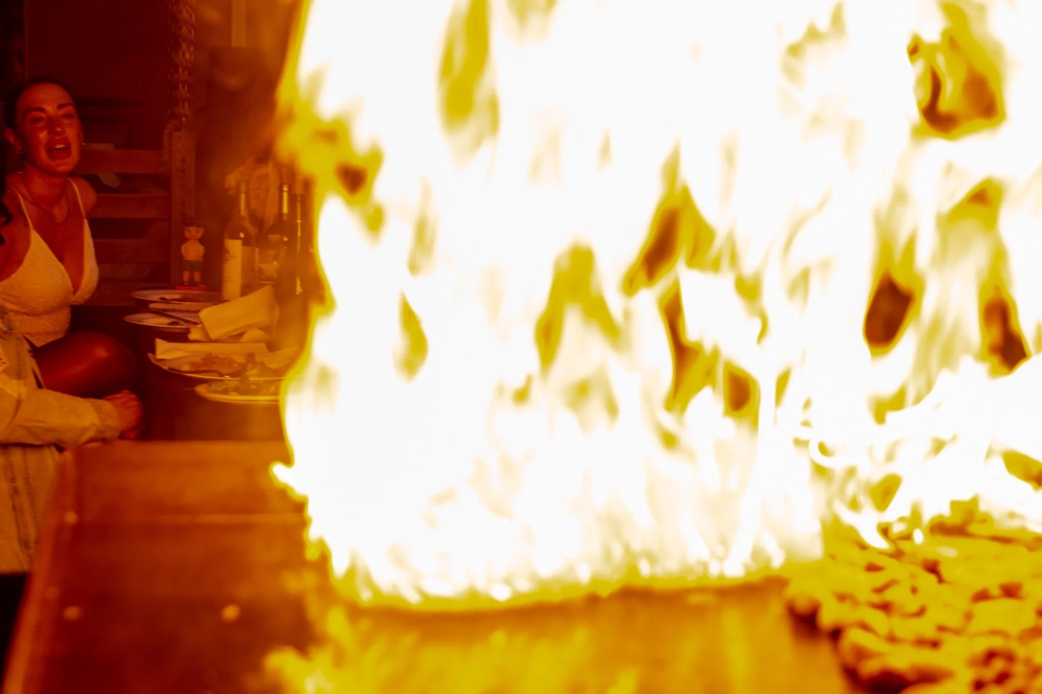 People gathered around a table with food, while large flames are in the foreground.