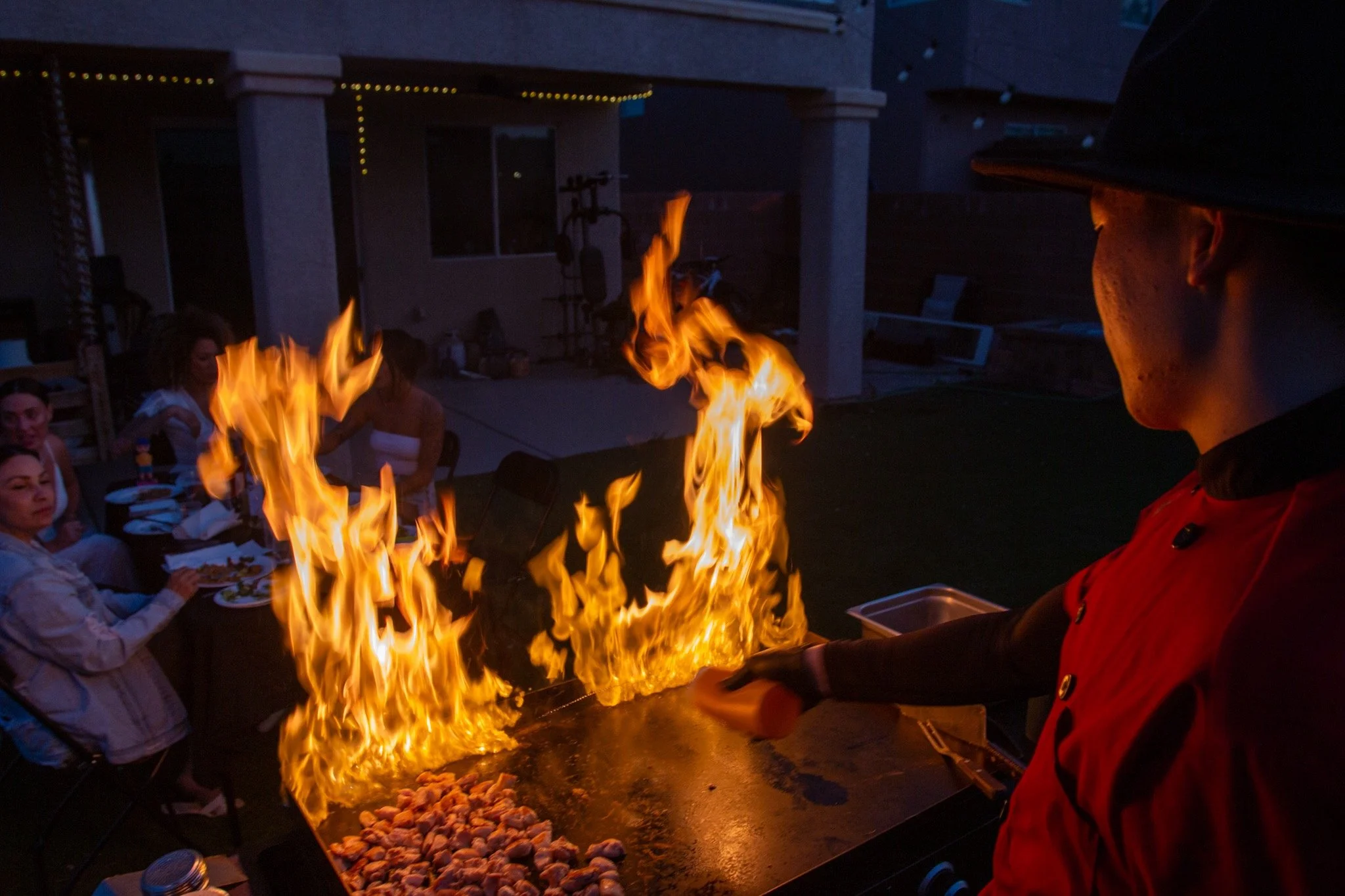 A chef in a red jacket and black hat cooks on a grill with large flames, while a group of people sit at a table nearby watching the cooking process at an outdoor gathering during evening.