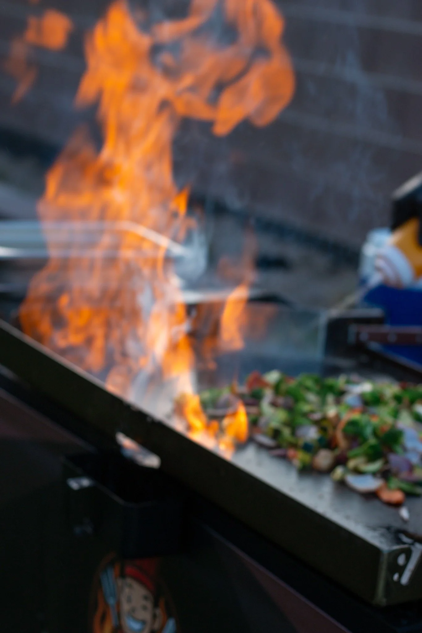 A flat-top grill with a small fire and smoke, cooking a mix of chopped vegetables.