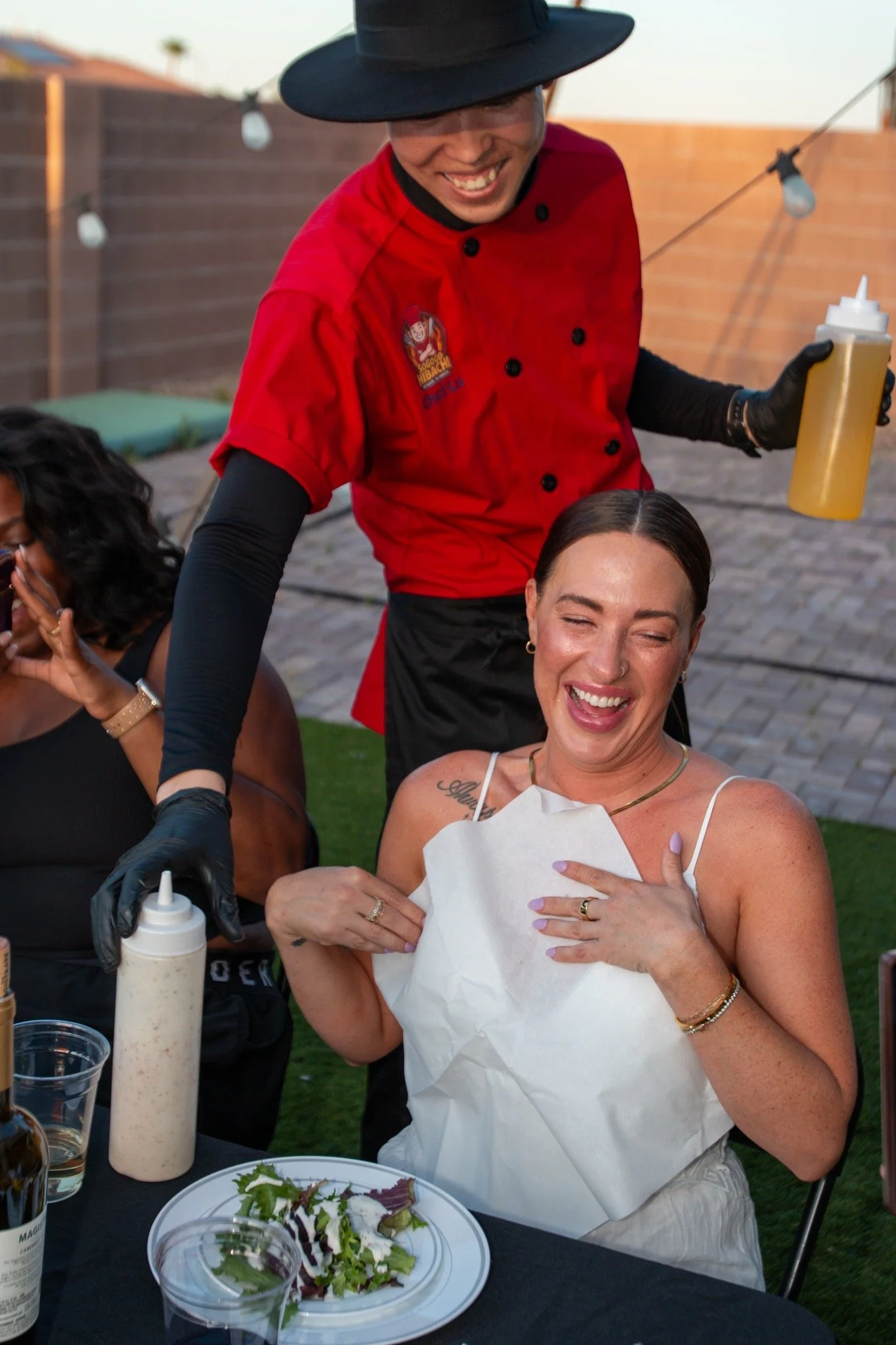 A woman laughing and smiling while receiving a picnic-style meal outdoors, with a server in a red uniform and black hat holding a squeeze bottle behind her.