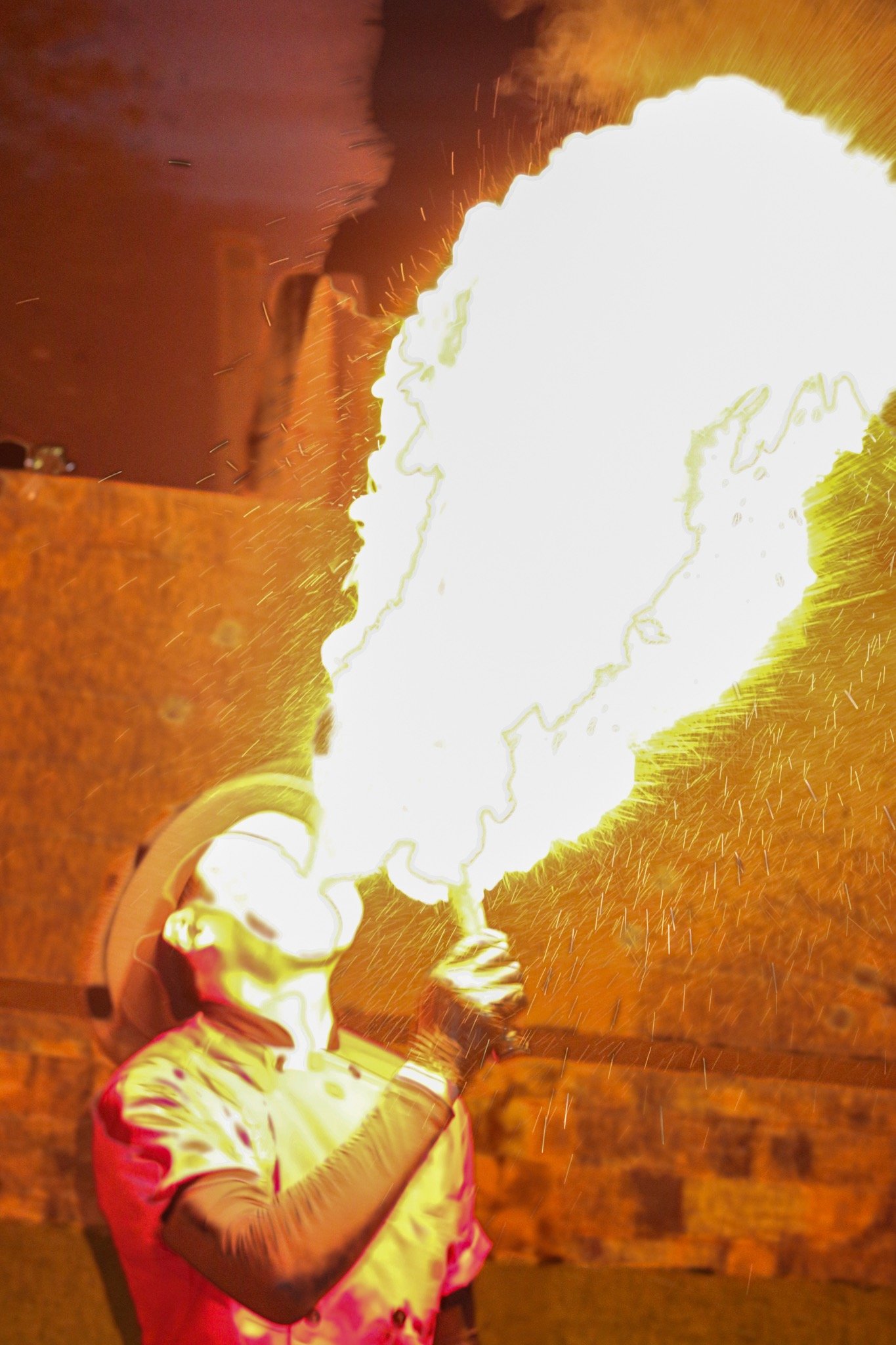 A person in protective gear is performing a fire-breathing act, creating a large fireball with sparks flying around.