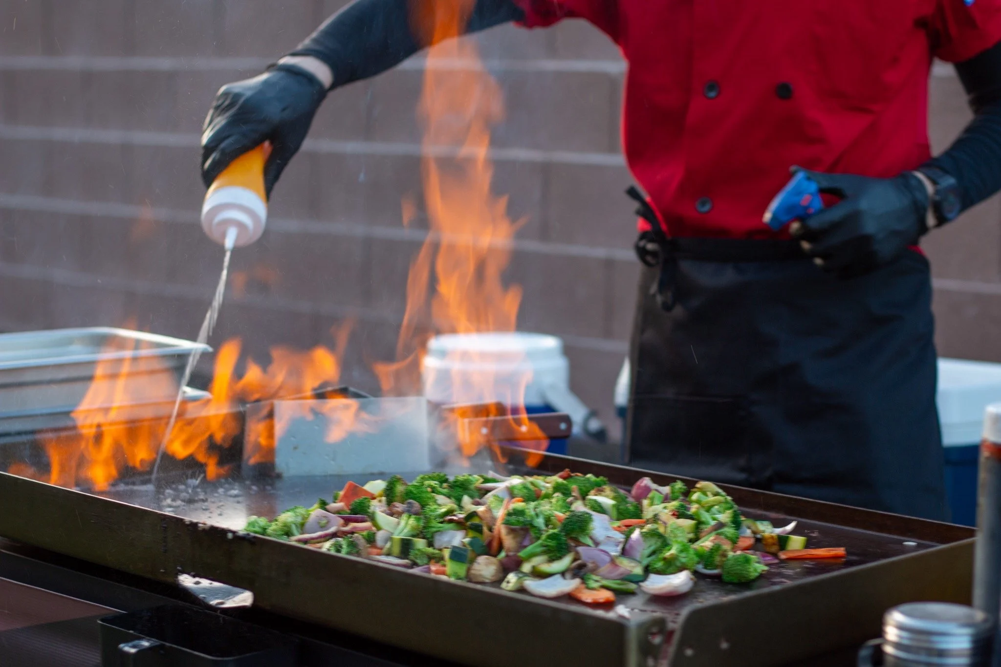 Chef wearing black gloves and a red apron cooking vegetables on a griddle with flames.