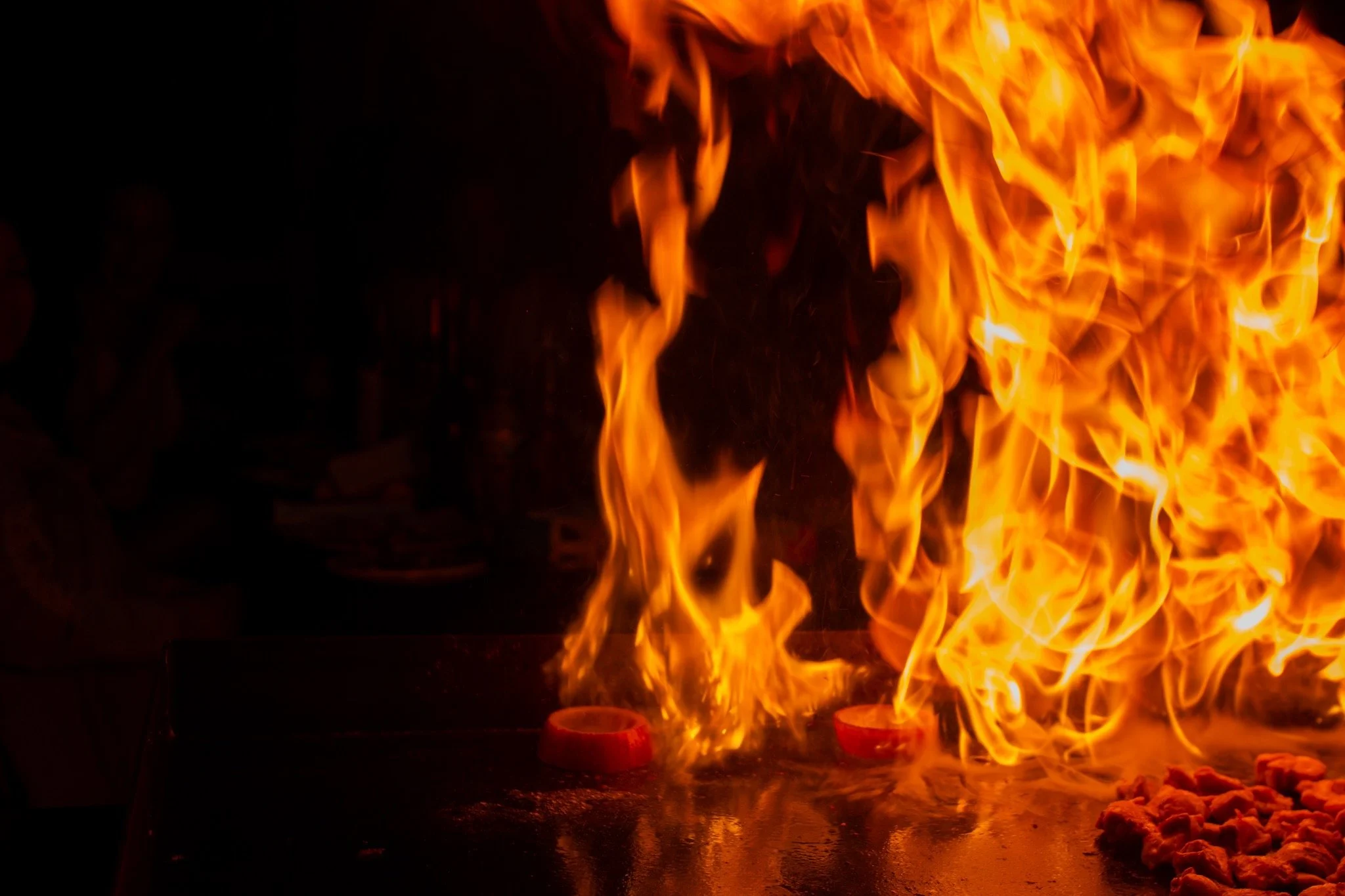 Large flames on a cooking surface with food, with a person faintly visible on the left side in a dark setting.