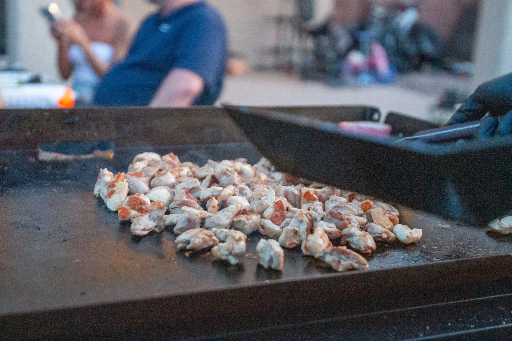 Chicken pieces on a grill or flat-top cooking surface, with people in the background.