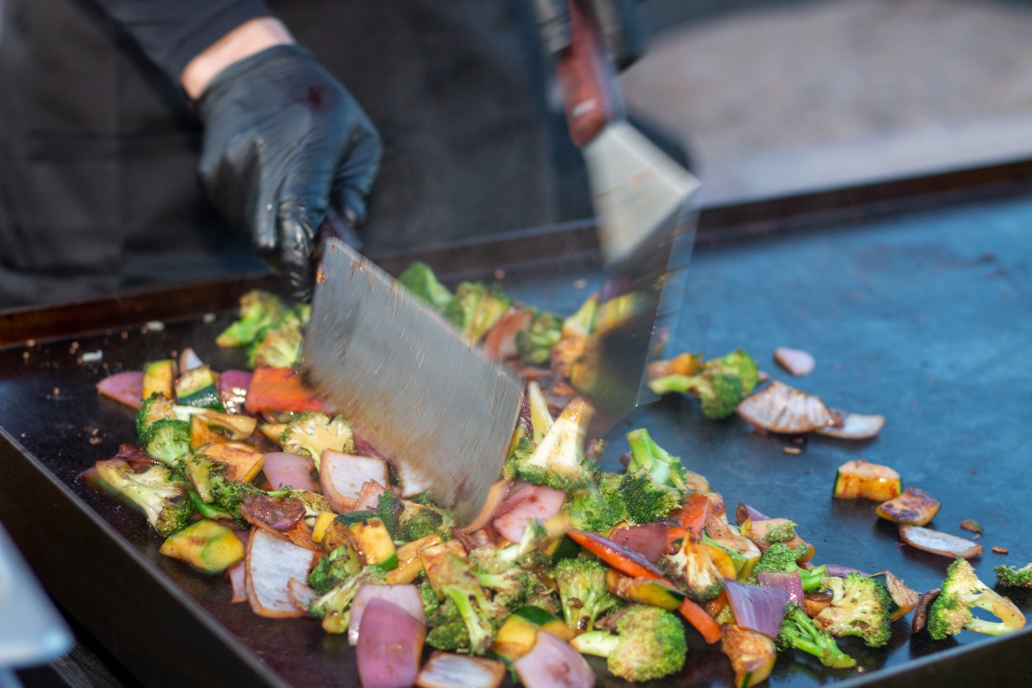 Person wearing black gloves and black apron cooking mixed vegetables on a flat-top grill, using a metal spatula.