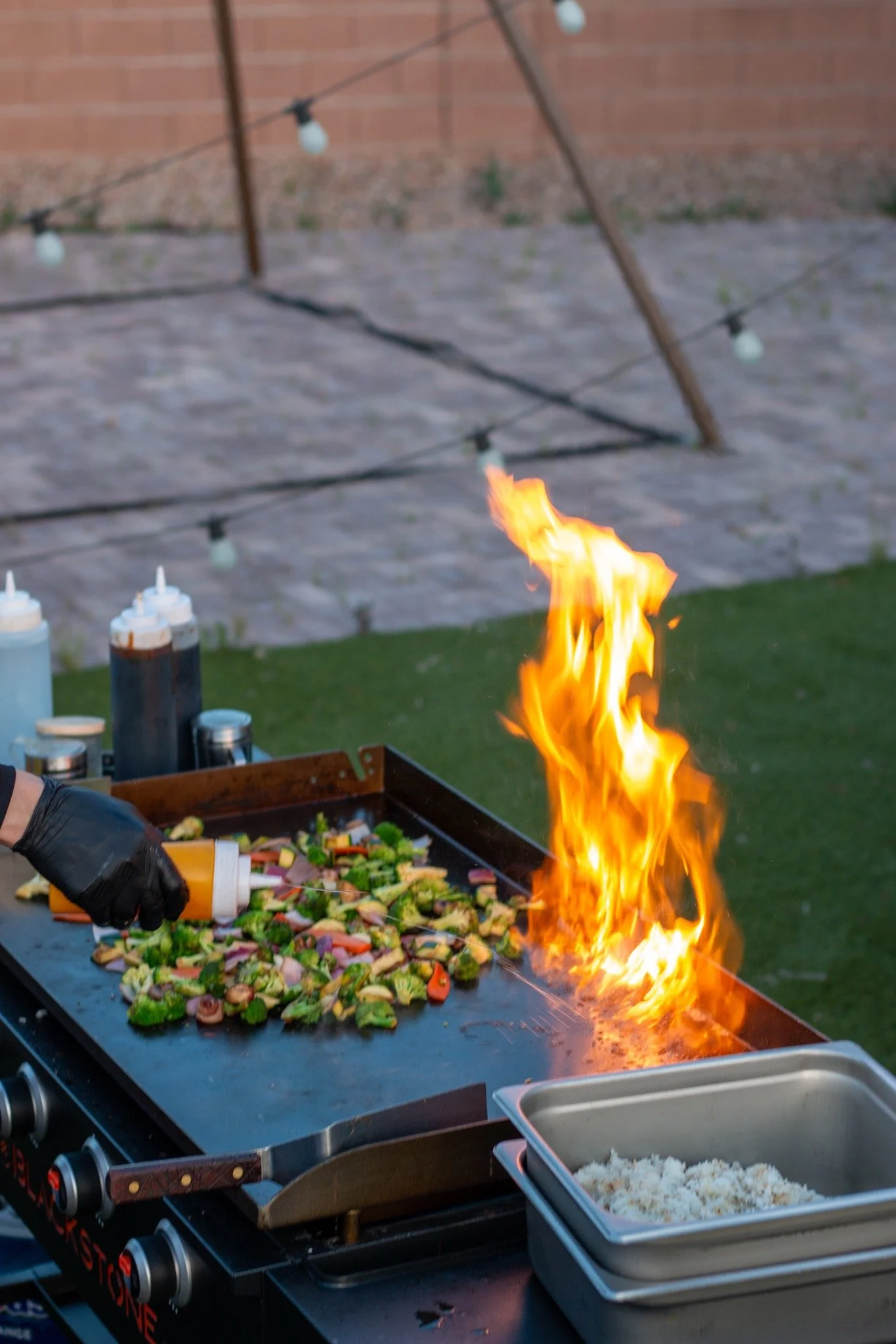 A chef wears black gloves and uses a squeeze bottle to add sauce to a colorful vegetable stir-fry on a grill, with flames rising from the food.