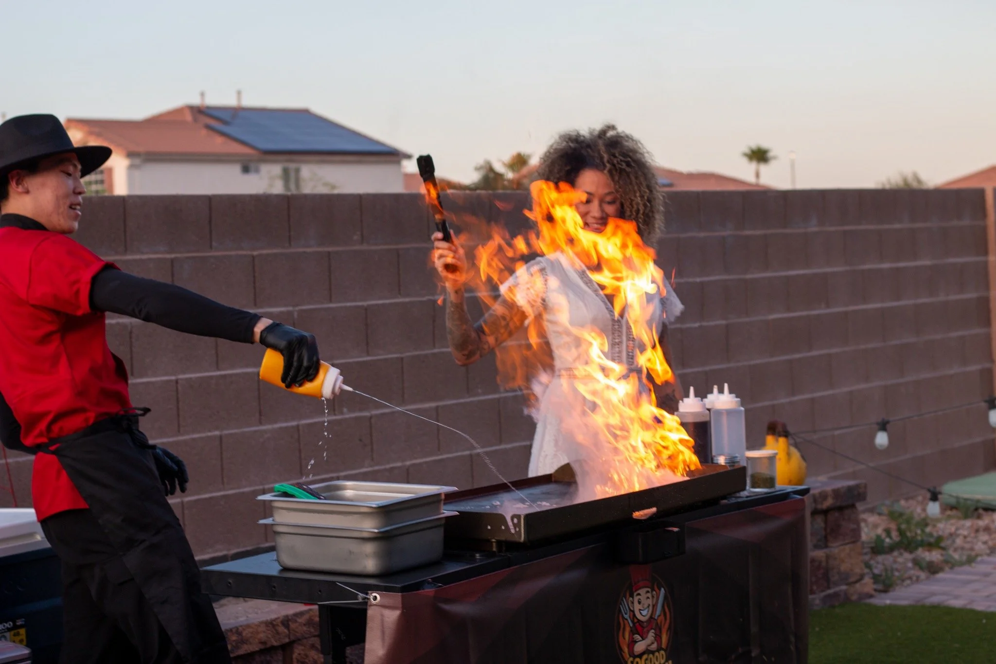 A woman is grilling with flames coming from her grill as she holds a spatula in the air, while a chef sprays fire retardant on the flames during an outdoor cooking event.