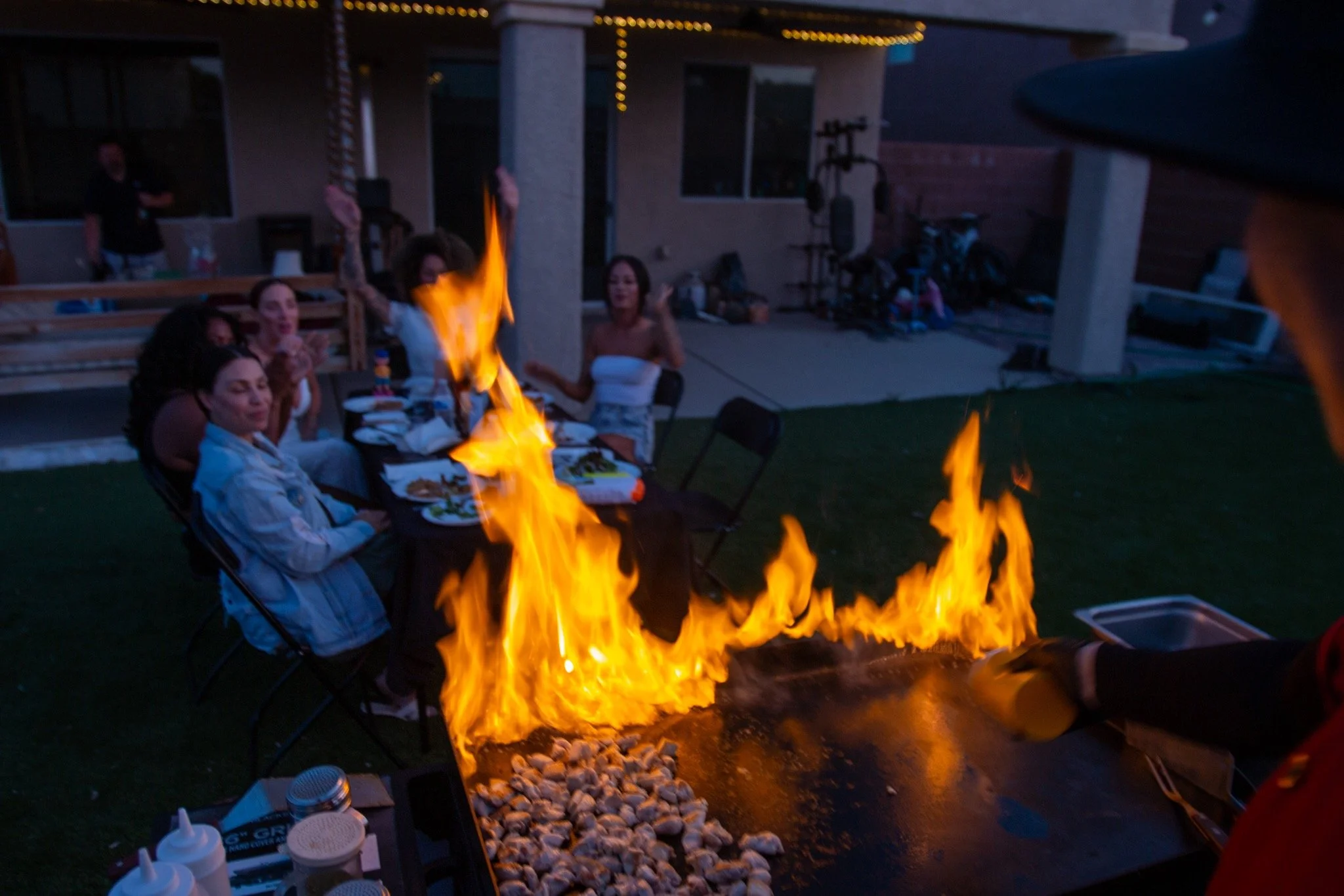 People sitting at a table outdoors during a barbecue with flames on a grill in front of them, some are raising their hands and smiling.