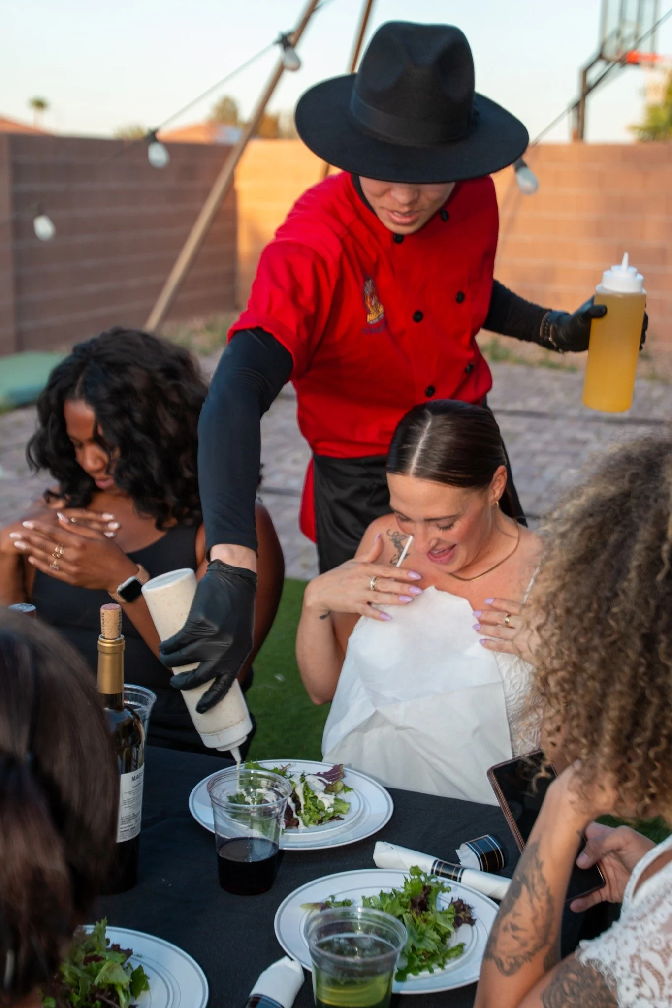 A woman in a white dress receiving her meal at an outdoor restaurant, a chef in a red uniform and black hat serving a plate of salad, and other women sitting around the table with plates of salad and drinks.