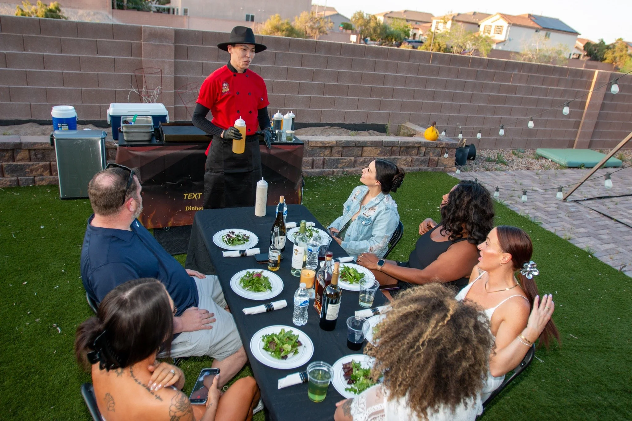 People sitting at a dinner table outdoors, listening to a chef prepare food. The chef is wearing a red shirt, black apron, and black hat. The table has plates of salad, bottles of wine, and drinks. There are string lights and a brick wall in the back