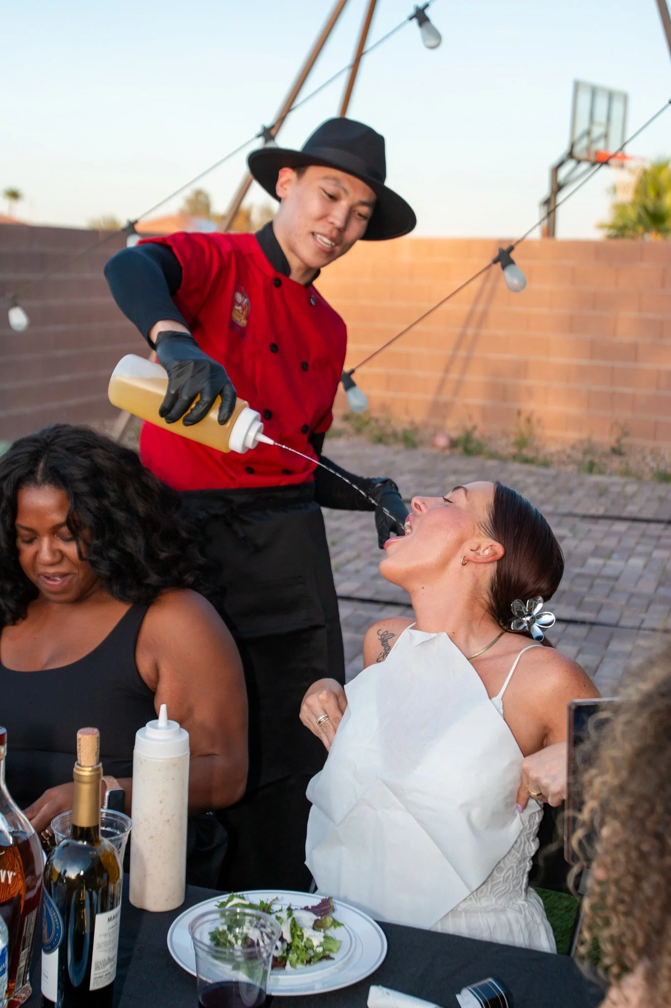 A woman at a dinner table opening her mouth wide as a man in a red chef uniform and black hat squirts sauce into her mouth during an outdoor gathering.