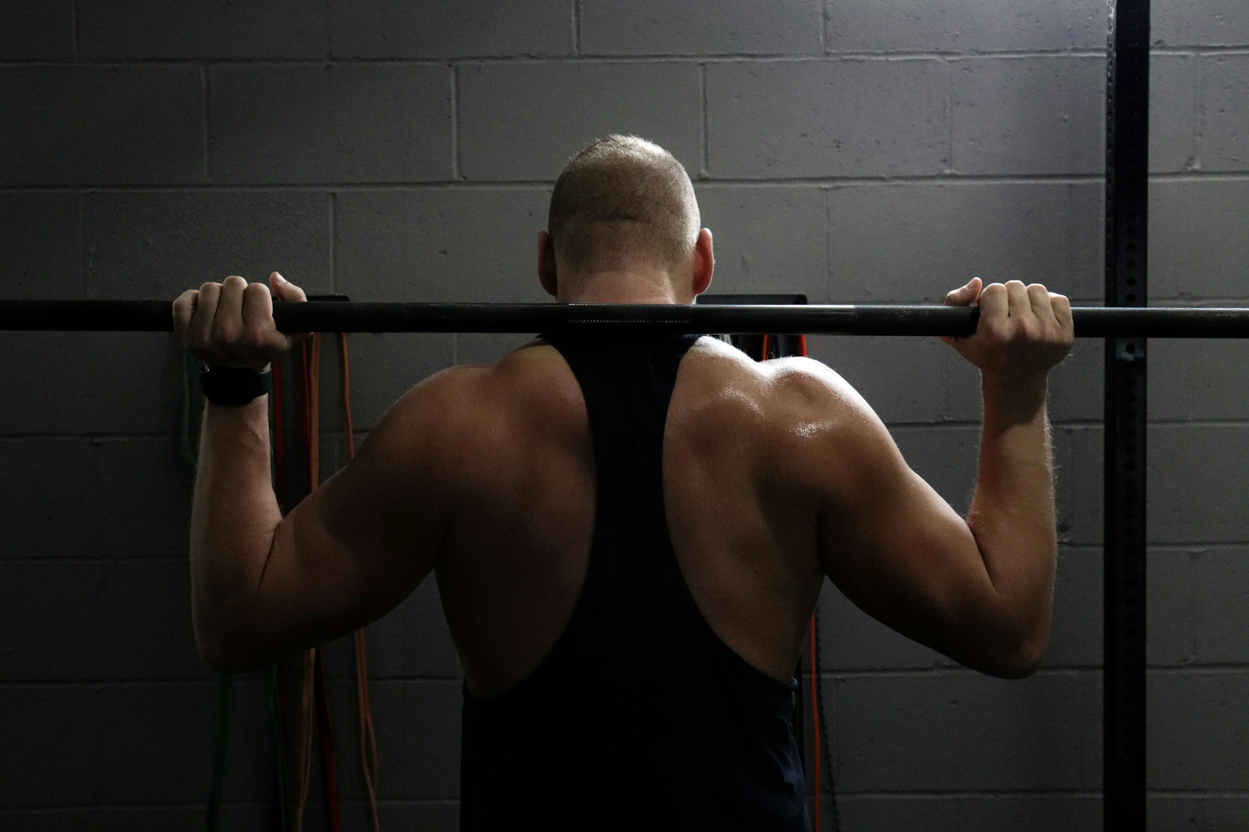 A muscular man with a shaved head is lifting a barbell at the gym, shown from behind.