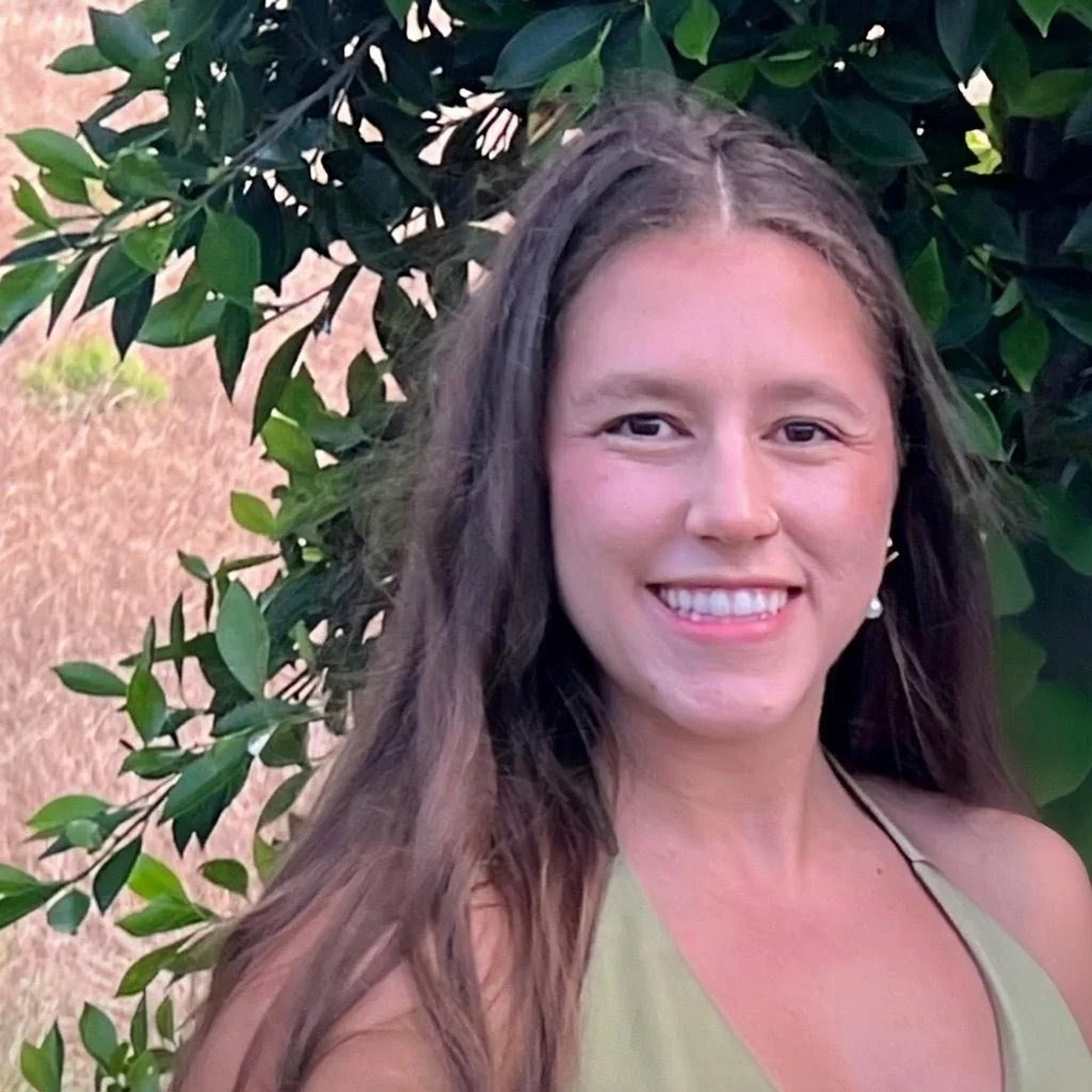 A woman with long brown hair smiling in front of green leafy shrubbery.
