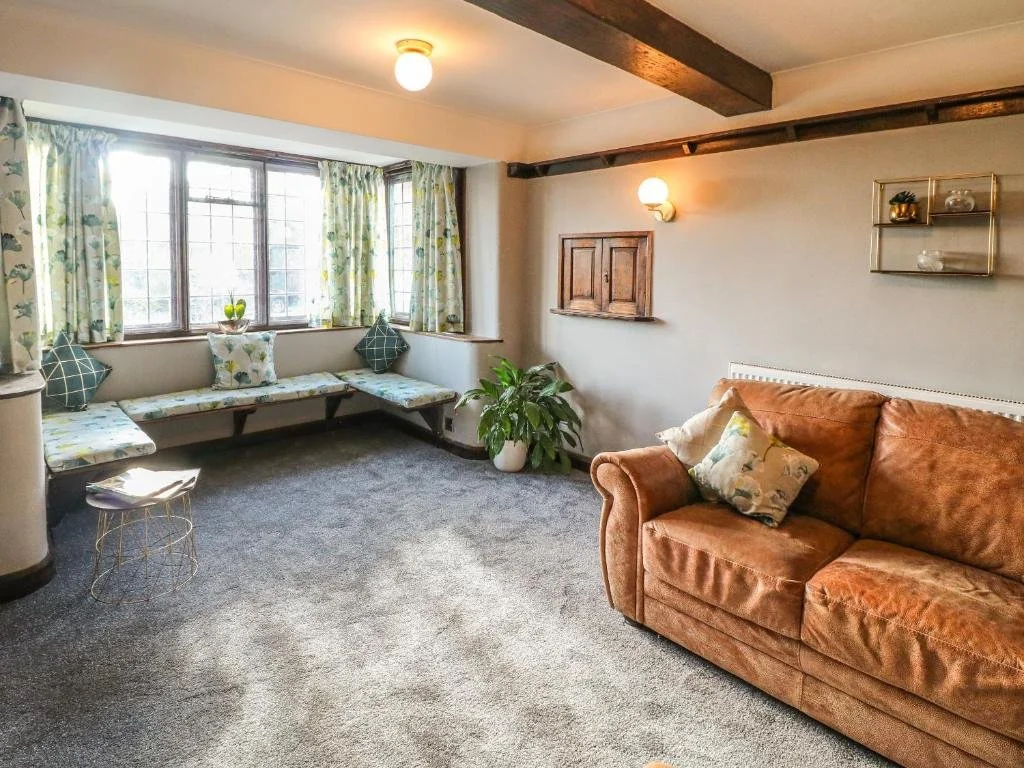 A cozy living room with a brown sofa, patterned pillows, a large window with floral curtains, built-in bench seating beneath the window, a potted plant, and decorative shelves on the wall.
