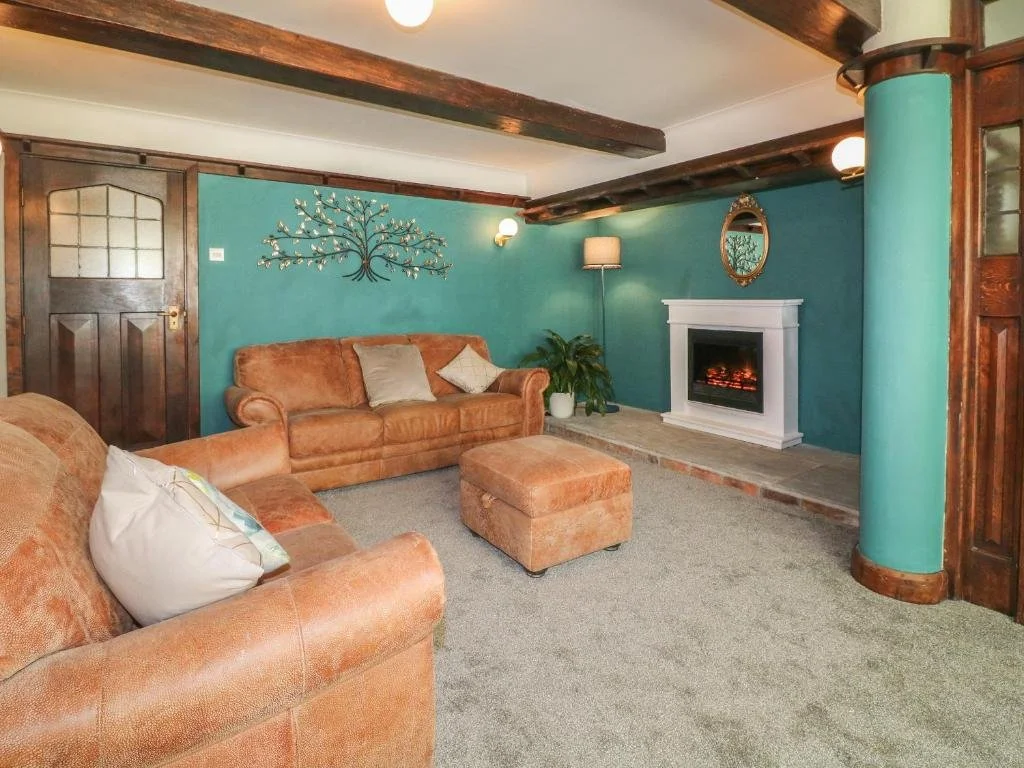 A cozy living room with teal walls, a fireplace with a white mantle, and two peach-colored sofas with pillows. There is a wall decoration of a metallic tree, a mirror above the fireplace, a tall floor lamp, and a potted plant. The ceiling has exposed