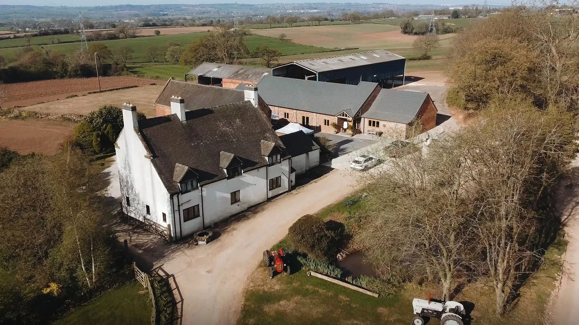 Aerial view of a farmstead with a white main house, several outbuildings, trees, a tractor, and expansive farmland in the background.