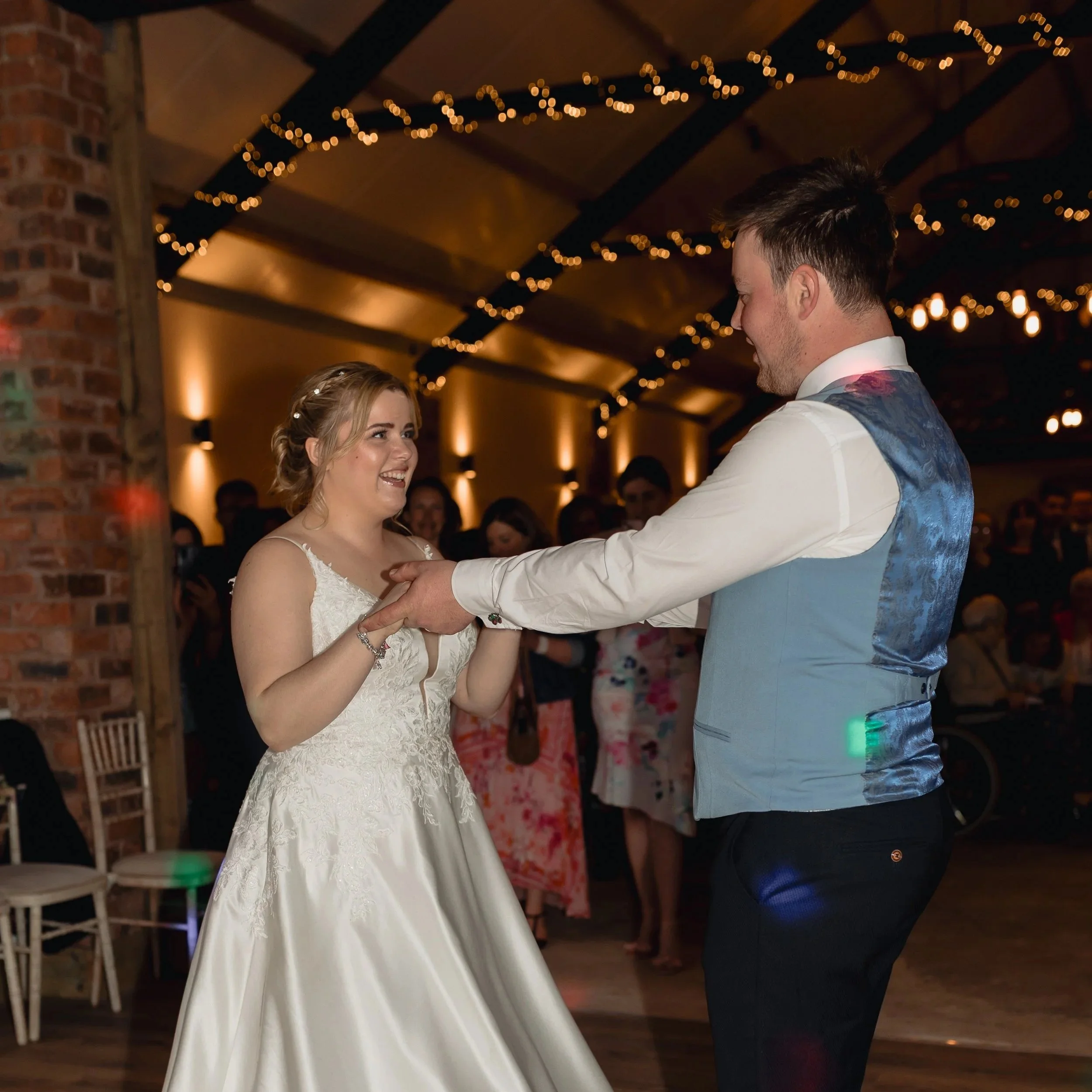 A bride and groom are dancing at their wedding reception under warm lighting with string lights overhead and guests in the background.