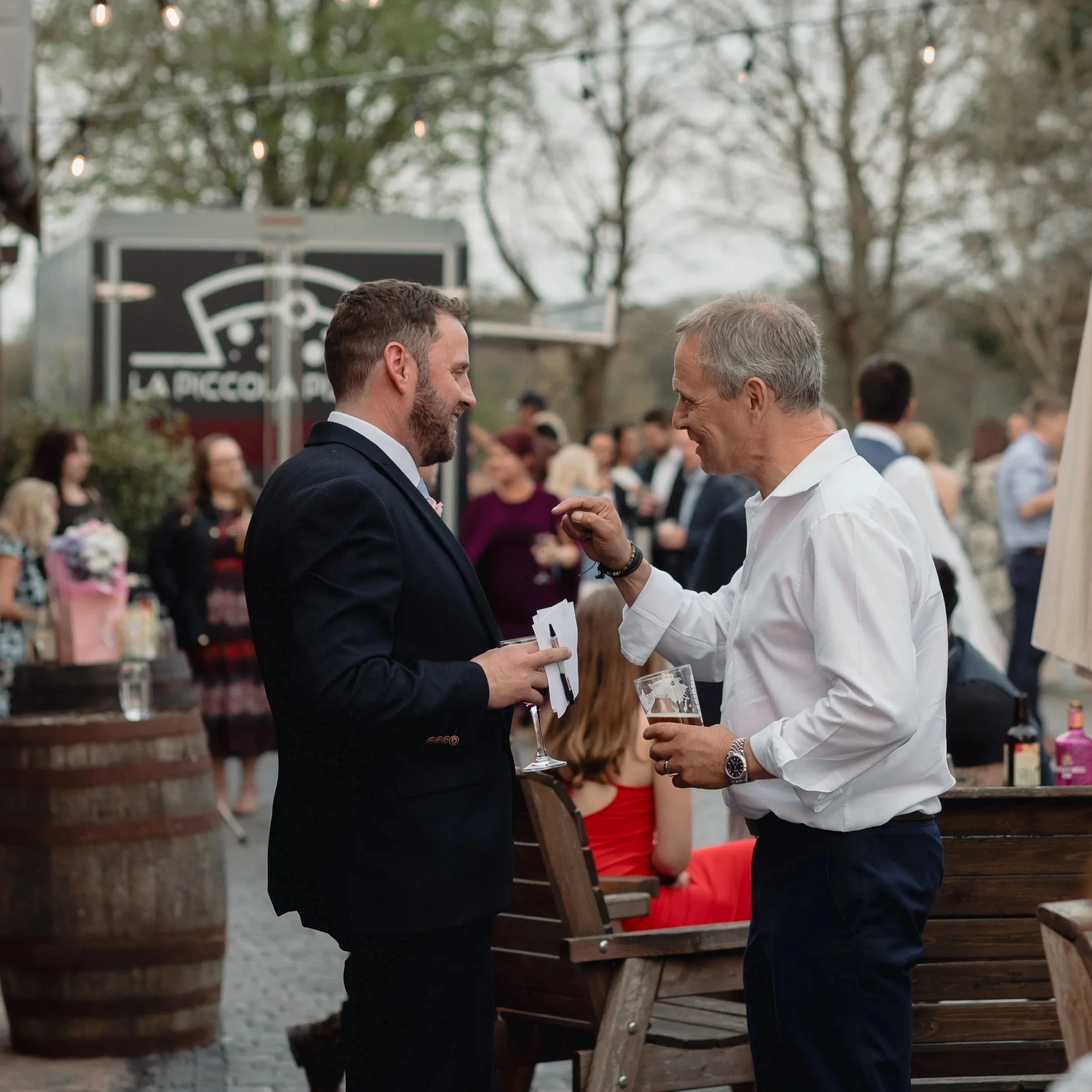 Two men in formal attire talking and smiling at an outdoor event, with people and trees in the background.
