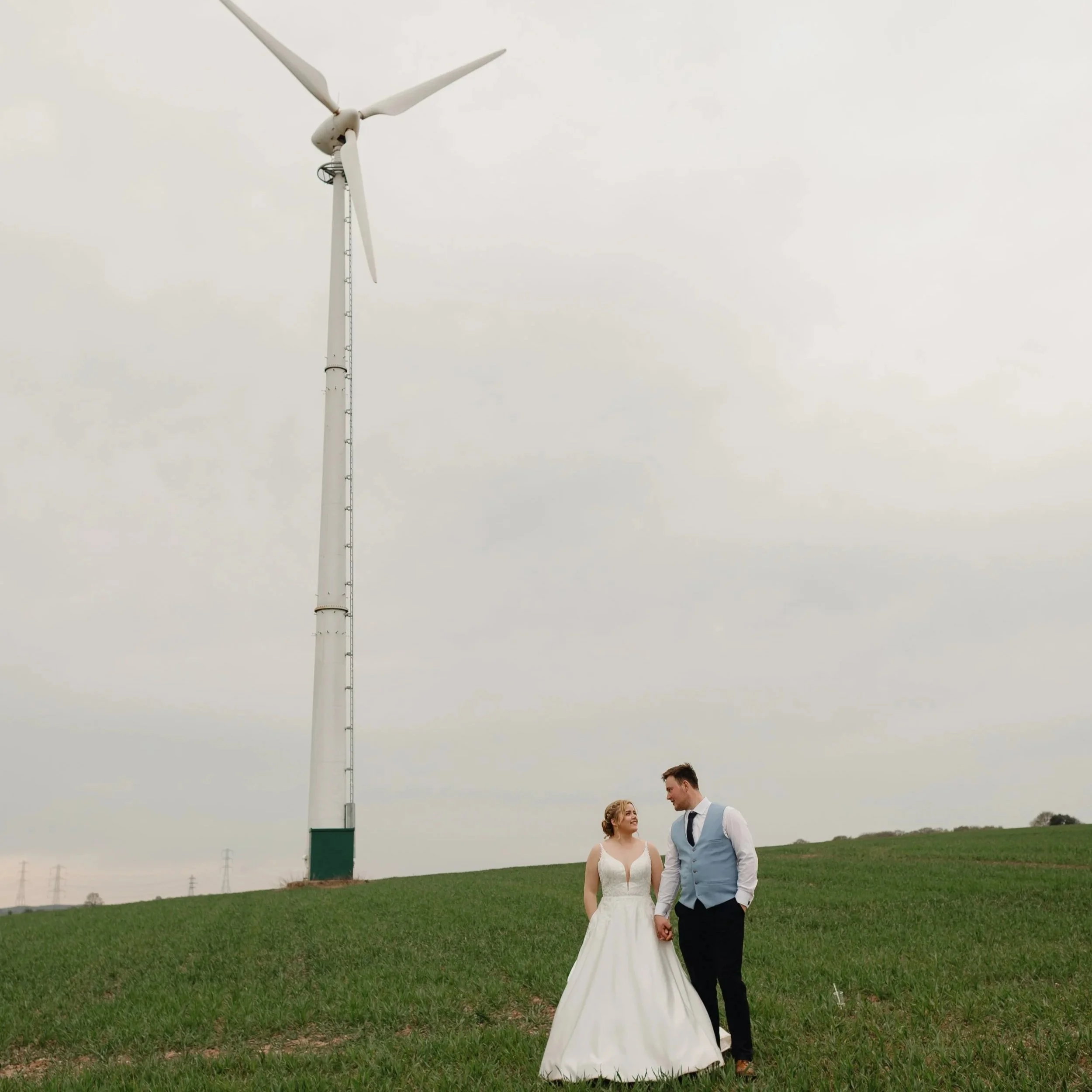 A bride and groom holding hands in a grassy field, with wind turbines in the background, on a cloudy day.