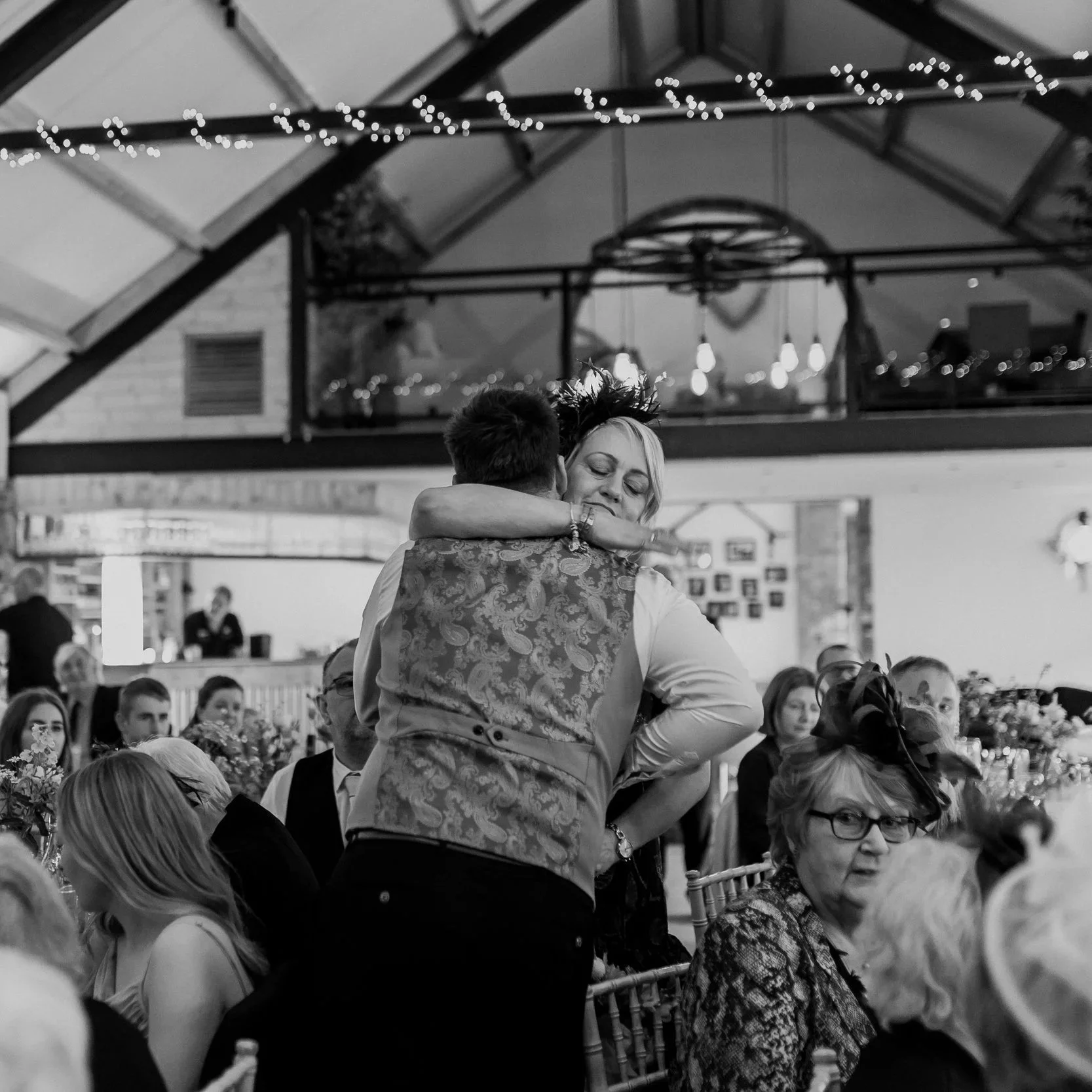 A black and white photo of a wedding reception showing a man and woman hugging. The woman is smiling and the man has his back to the camera. They are surrounded by seated guests, with a decorated setting including string lights overhead.