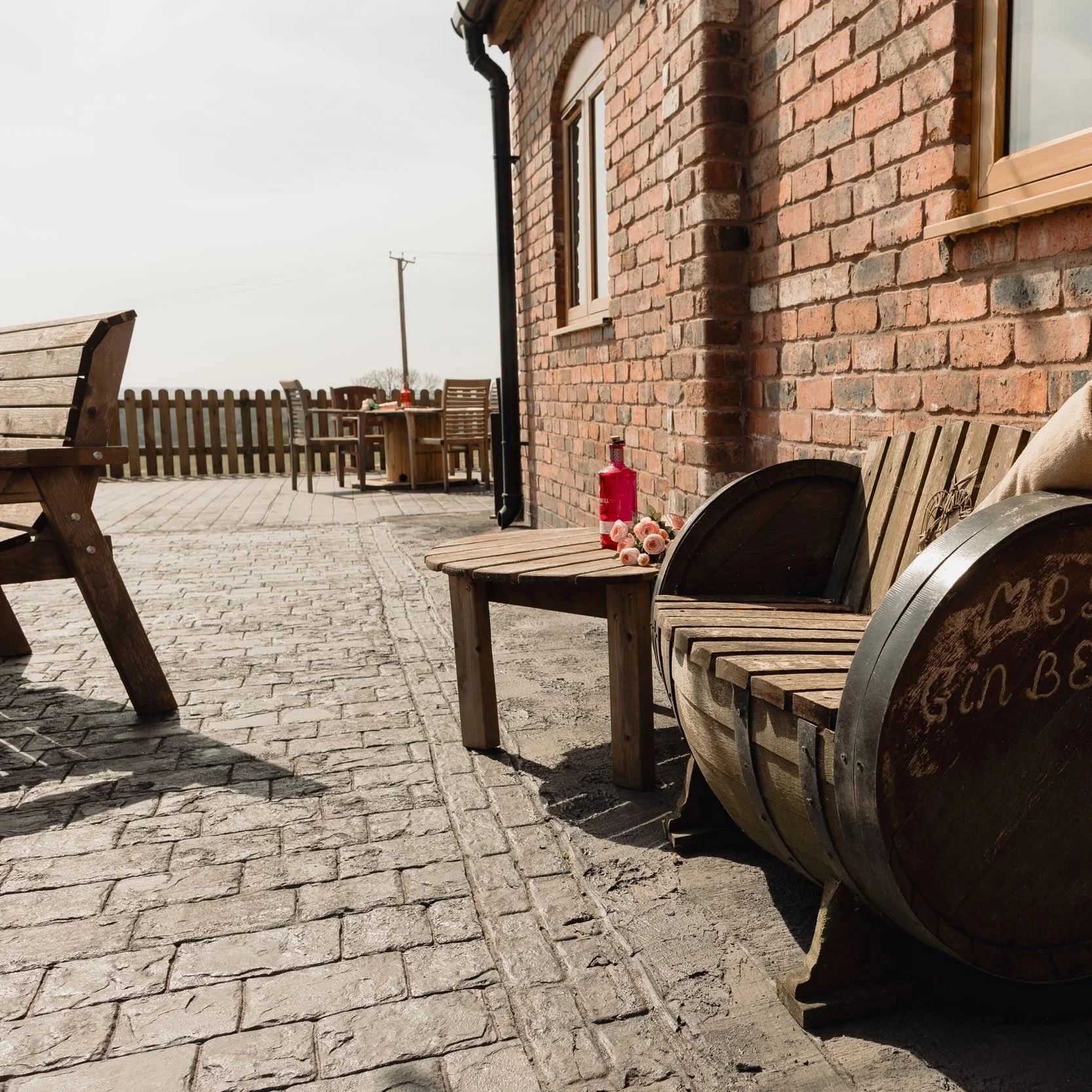 Outdoor patio with brick wall, wooden benches, small table with flowers, wine bottle, and a red fire extinguisher, on a sunny day.