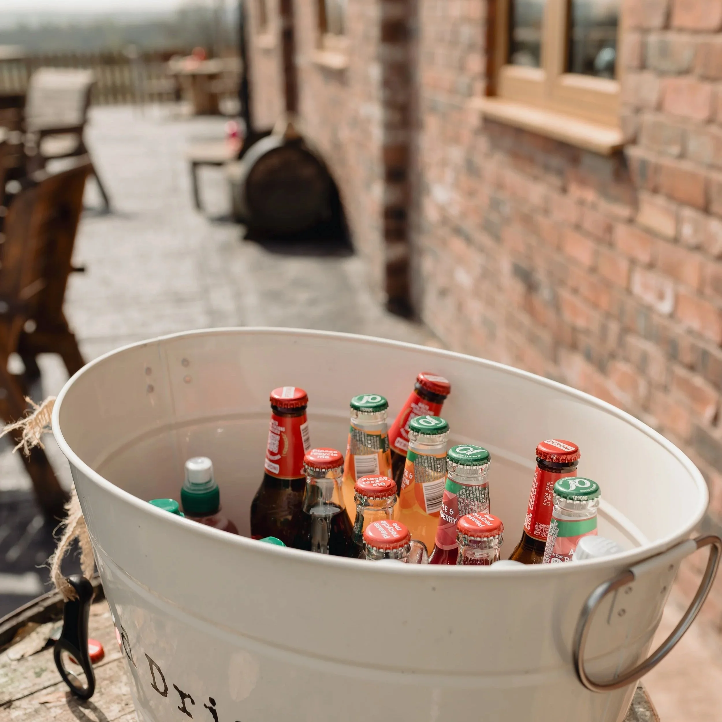 White bucket filled with small glass bottles with red and green caps, placed outside on a wooden surface near a brick building.