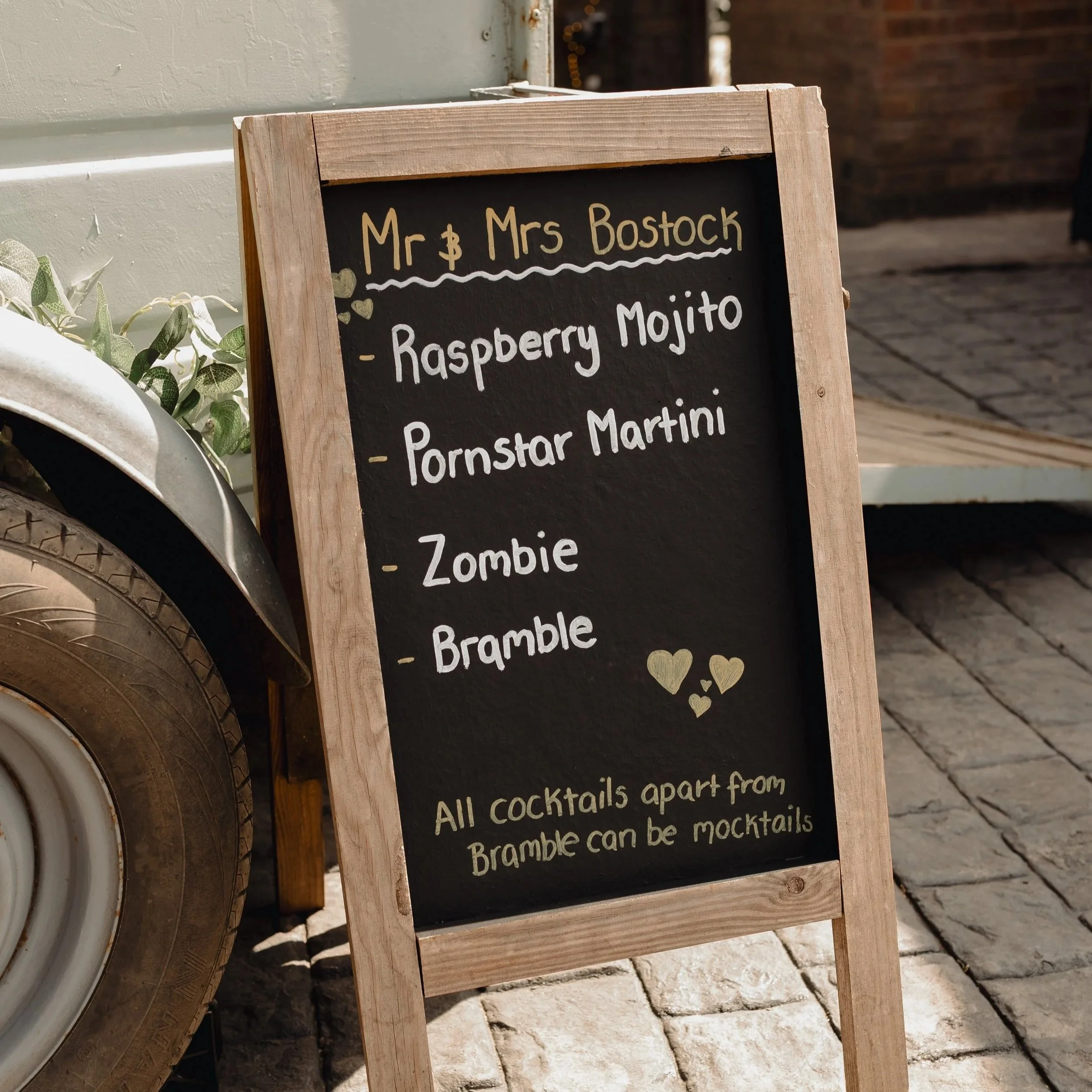 A black chalkboard sign with a light wooden frame listing cocktails available outside a venue. The sign reads: 'Mr & Mrs Bostock, Raspberry Mojito, Pornstar Martini, Zombie, Bramble' with decorative hearts and wavy lines, leaning against a wall next 
