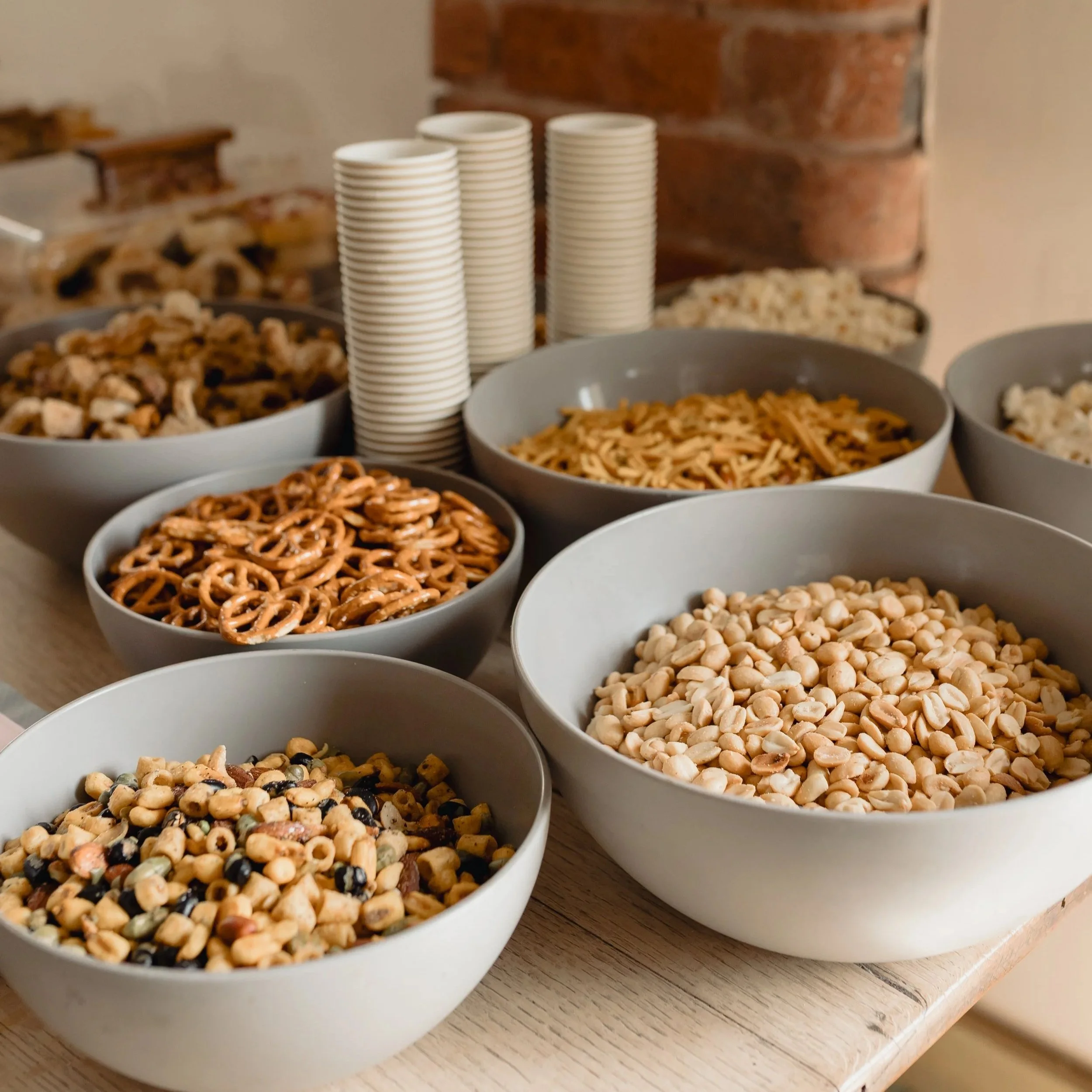 Bowl of on-the-go snack foods, including pretzels, chickpeas, and trail mix, with stacks of paper cups and bowls of trail mix on a wooden surface.